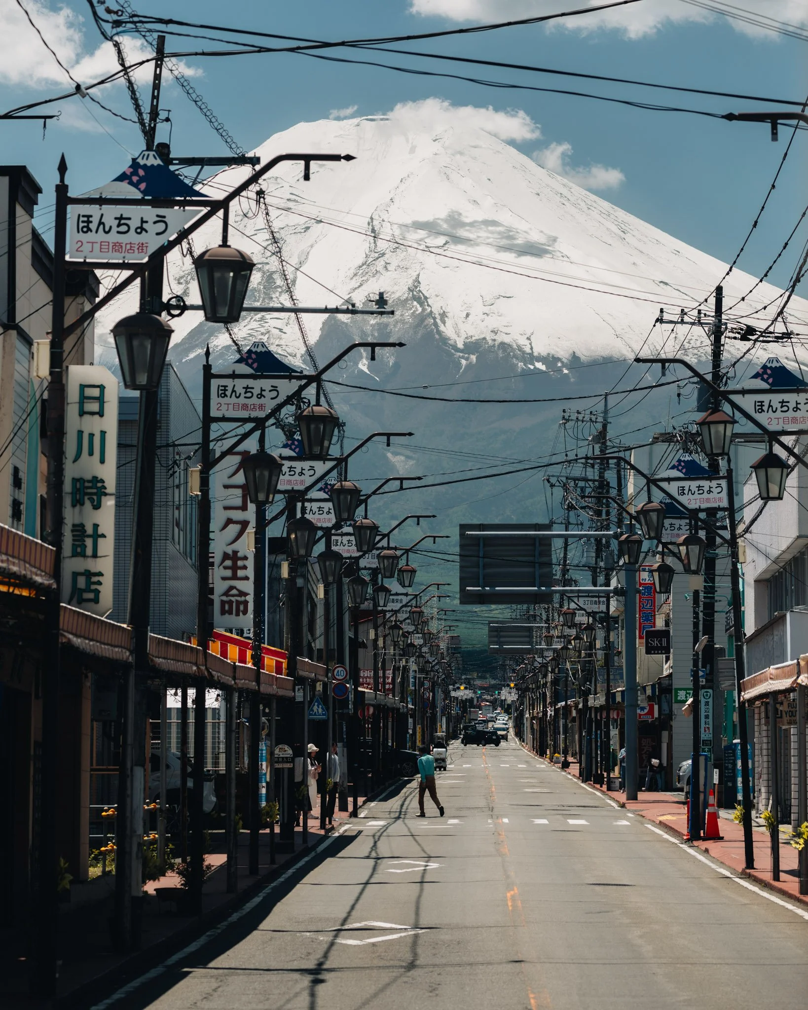 A street scene in Japan with Mount Fuji in the background. The street is lined with signs, street lamps, and power lines. There are a few pedestrians walking on the sidewalk and some cars on the road.