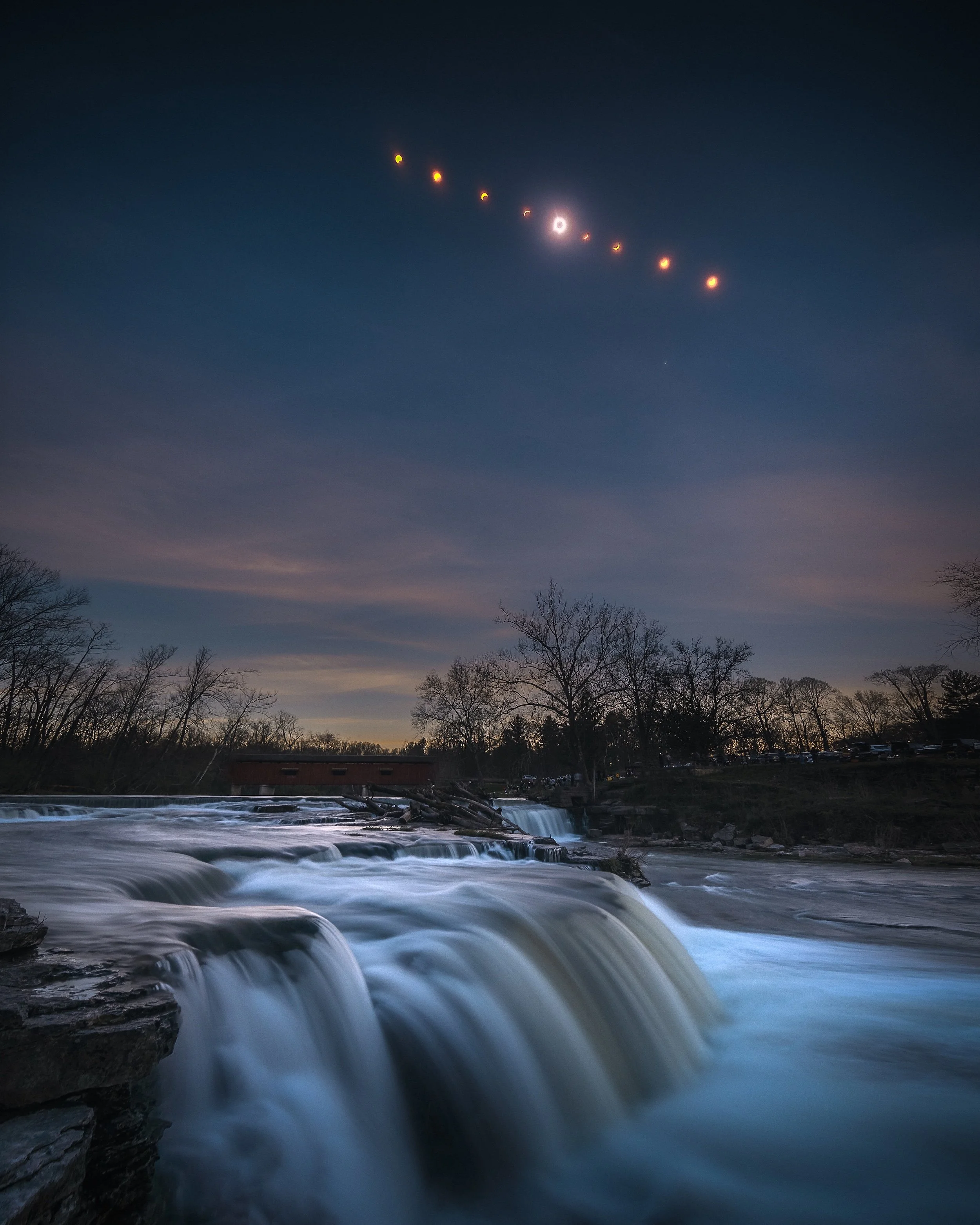 A river flows over small waterfalls with trees in the background and a cloudy sky during dusk.