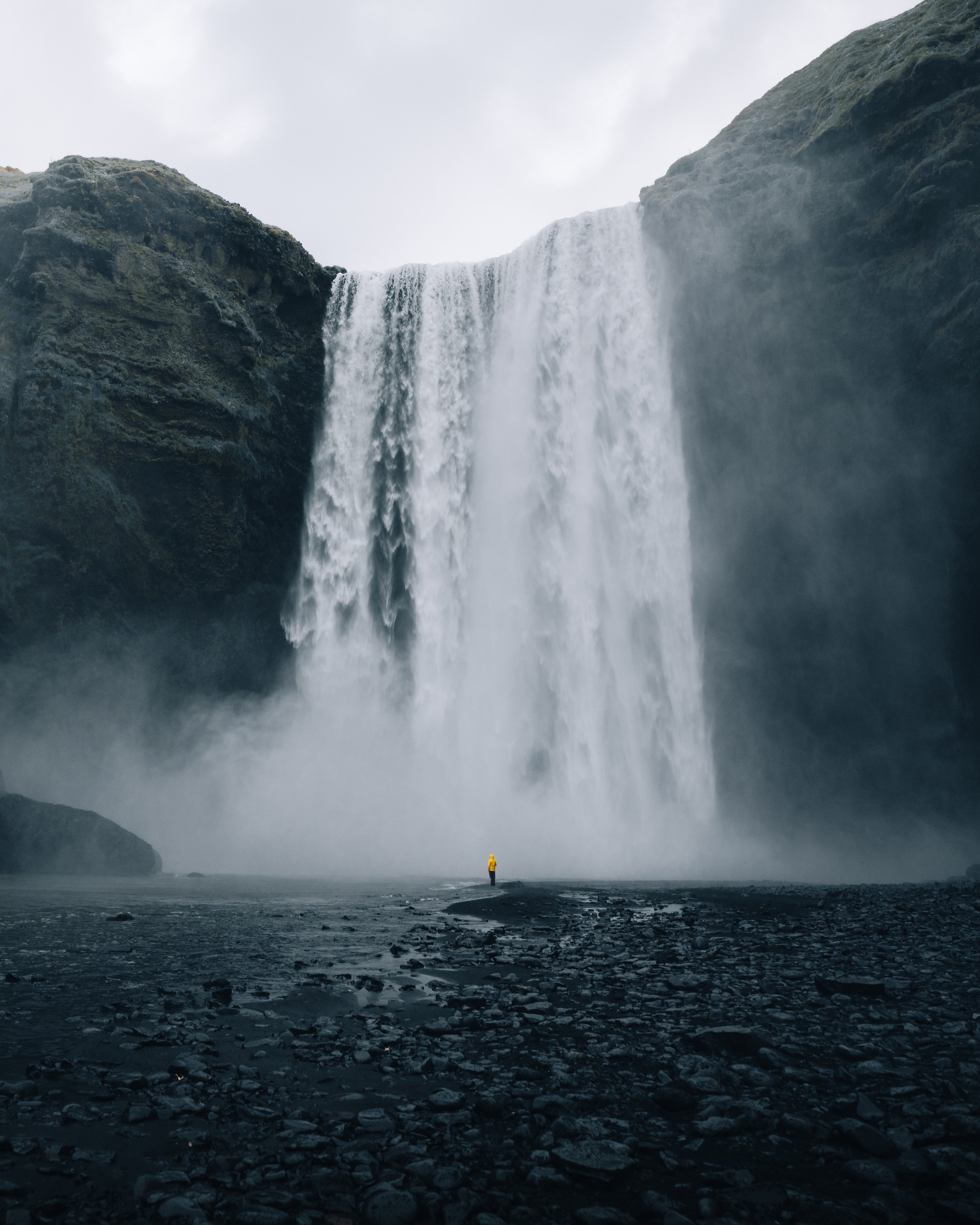 A person in a yellow jacket standing on rocky ground near the water, with a large waterfall cascading down from cliffs into the misty basin below.