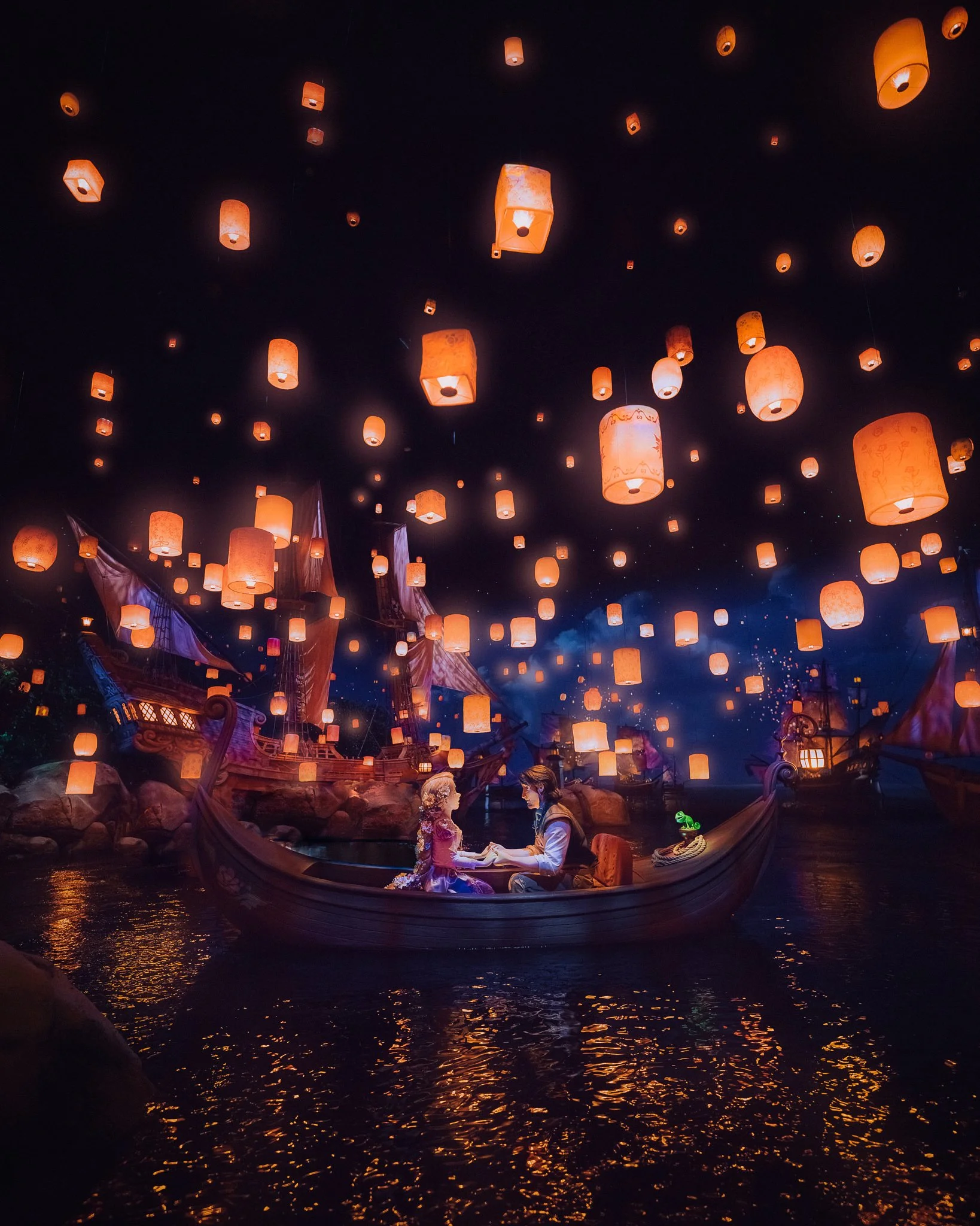 A boat with two people, a man and a woman dressed in costumes, holding hands on a river at night. There are many orange lanterns floating in the dark sky above them, creating a magical scene.