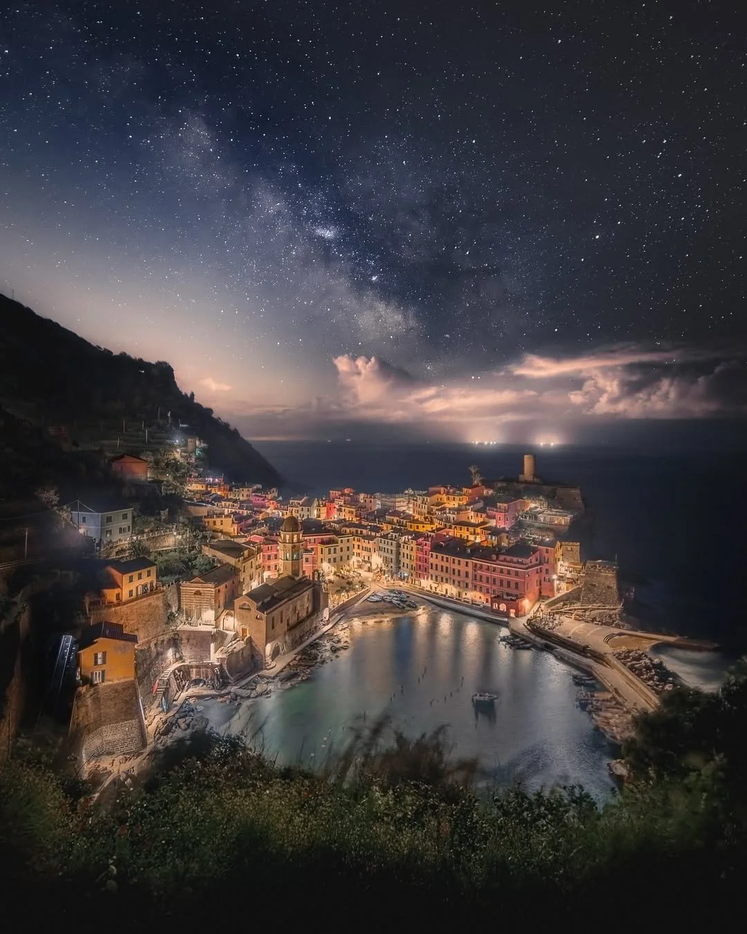 Night view of a coastal Italian village with colorful buildings, a small harbor, and a hillside under a starry sky with the Milky Way galaxy.