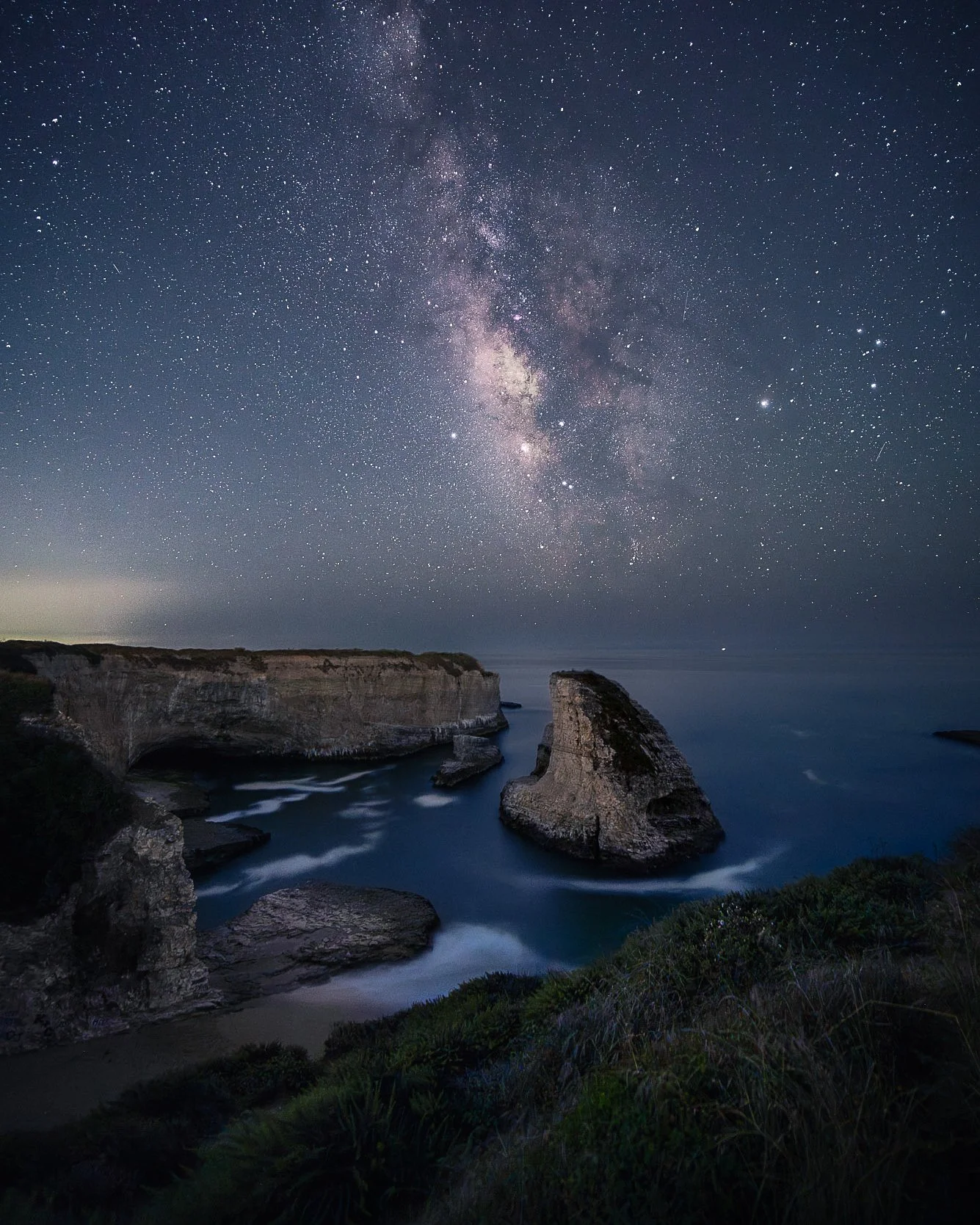 Nighttime view of a star-filled sky and the Milky Way galaxy above a coastal landscape with cliffs and ocean waves.