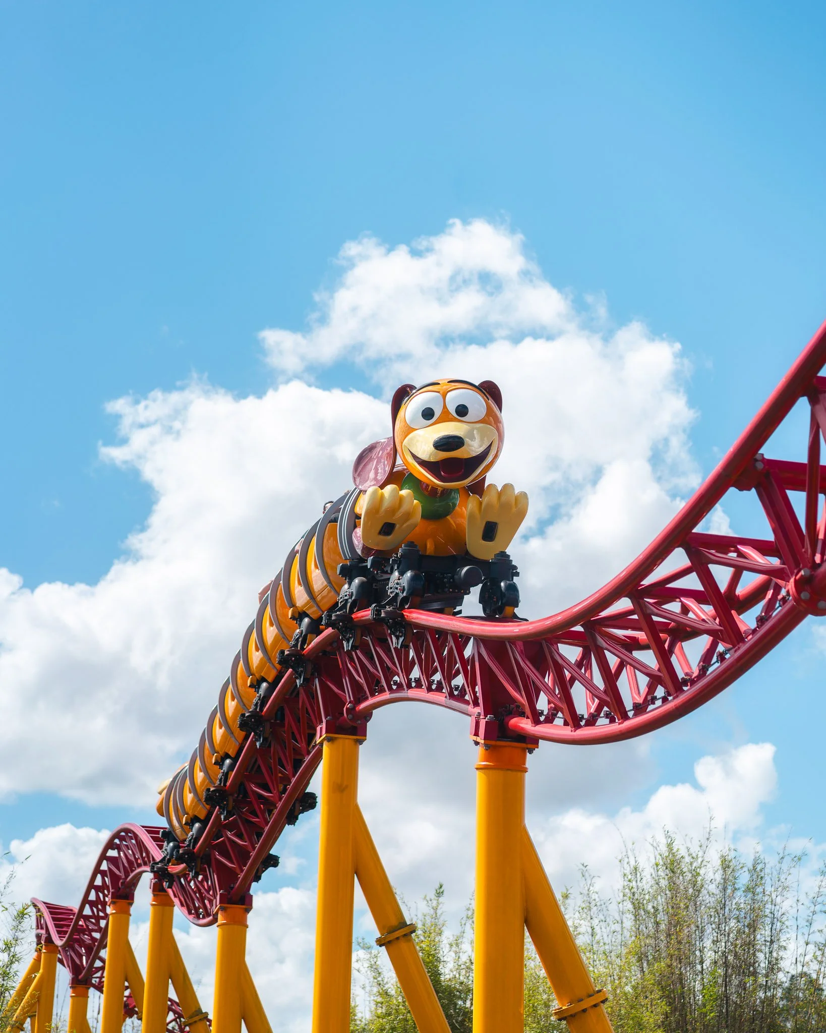 A roller coaster featuring a cartoon dog character with large eyes and a happy expression, riding on a bright red track with yellow supports, set against a blue sky with scattered white clouds and some trees in the background.