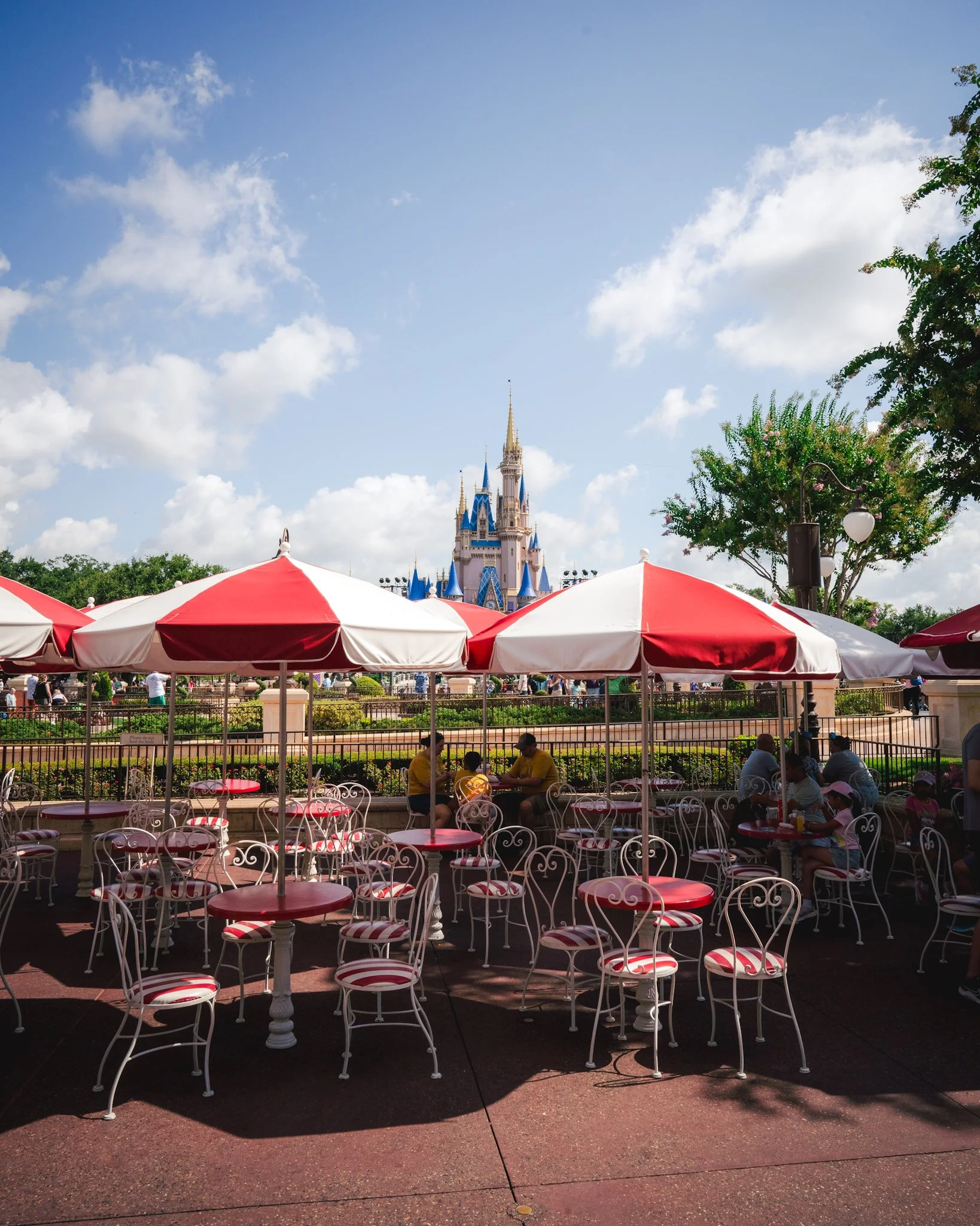 Disney castle in the distance under a blue sky with clouds, surrounded by trees. In the foreground, there are red and white umbrellas over seating areas with white metal chairs, and people sitting and walking around.
