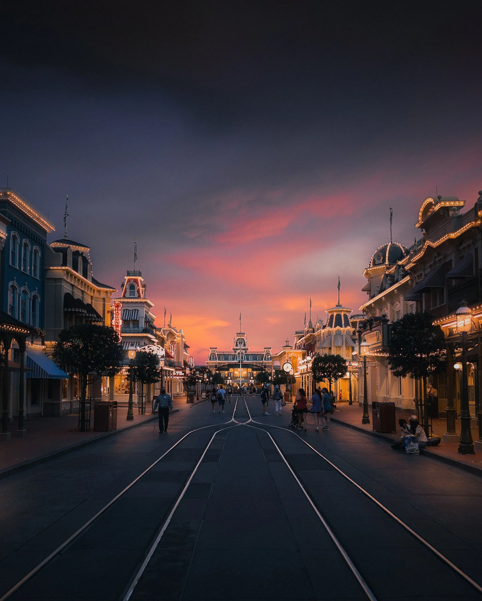 A street view of a Disney theme park at sunset with historic-style buildings illuminated with lights and people walking along the street, some pushing strollers, and tram tracks running down the middle of the street.