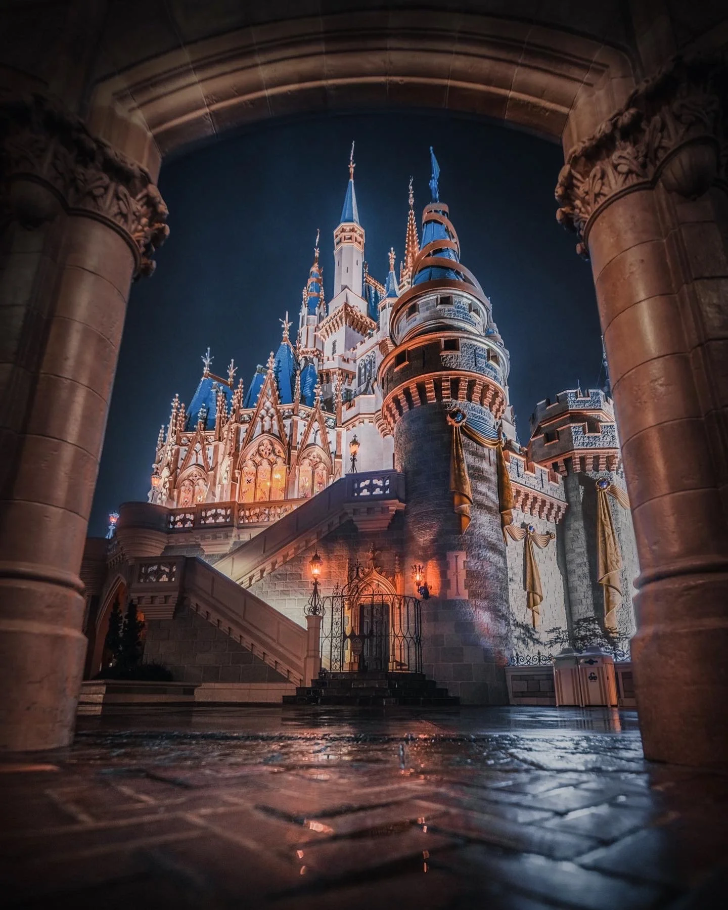 Nighttime view of a Disney castle illuminated with warm lights, viewed through an arched stone entrance. The castle has blue turrets and tall spires, with ornate architectural details.
