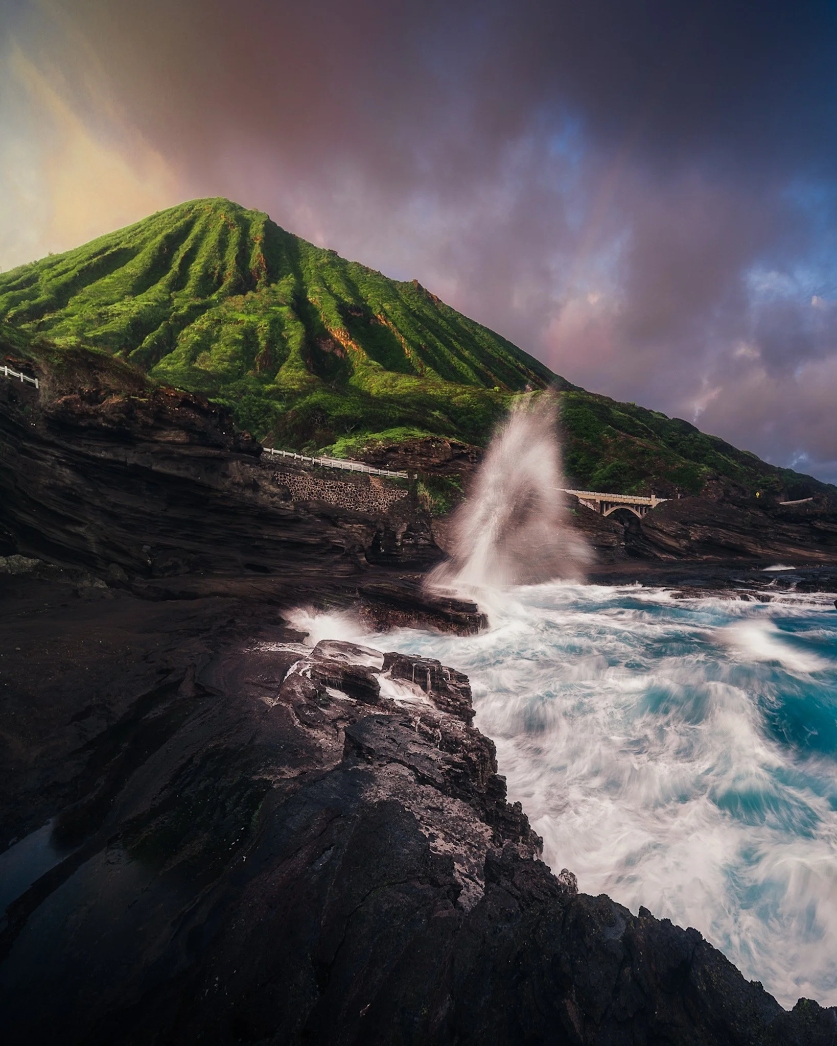A lush green mountain with steep ridges, a dark rocky shoreline, and ocean waves crashing against the rocks, with a cloudy sky overhead.