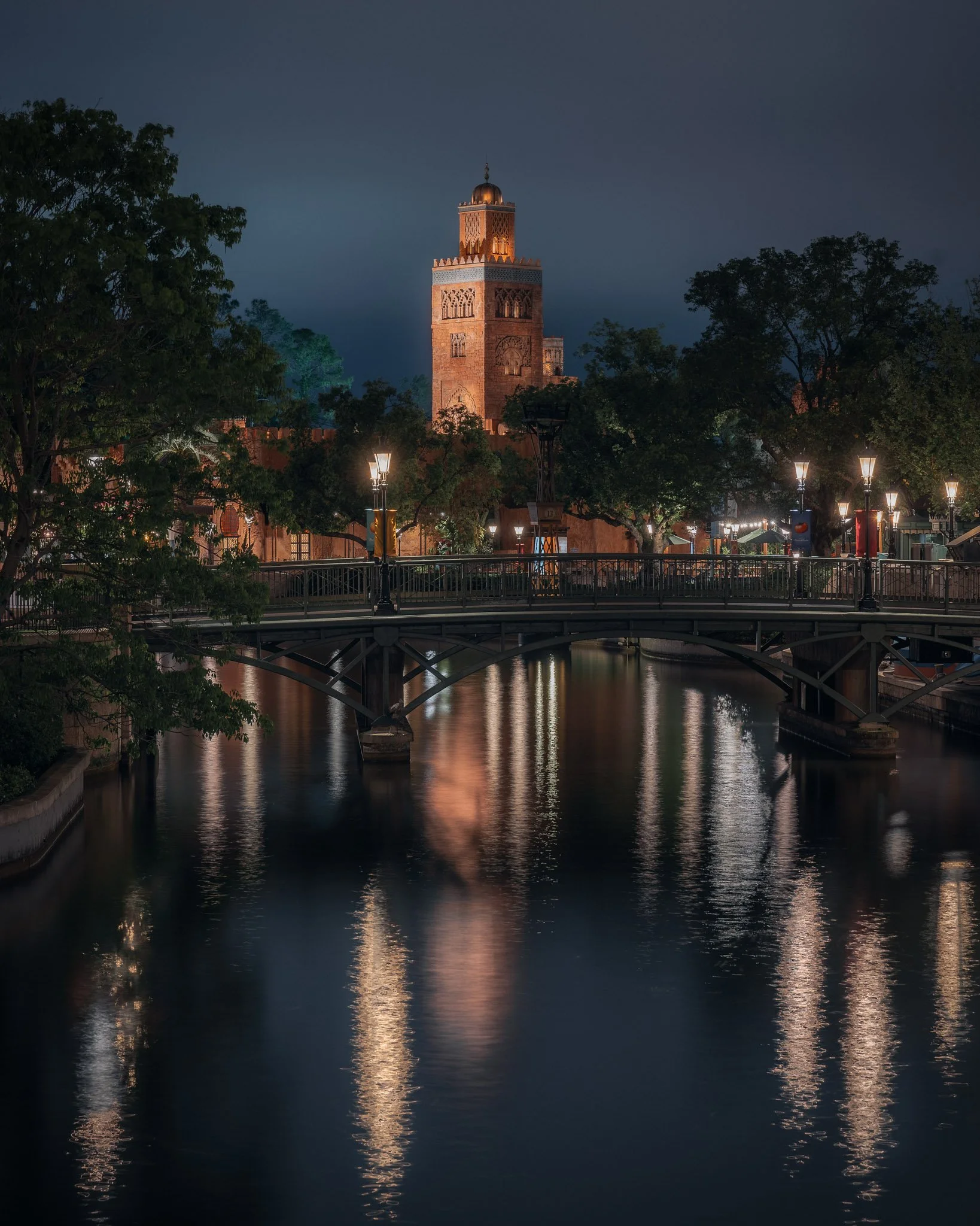 Night view of a historic mosque tower with intricate designs, illuminated and reflected in a river, with trees and street lamps in the foreground.