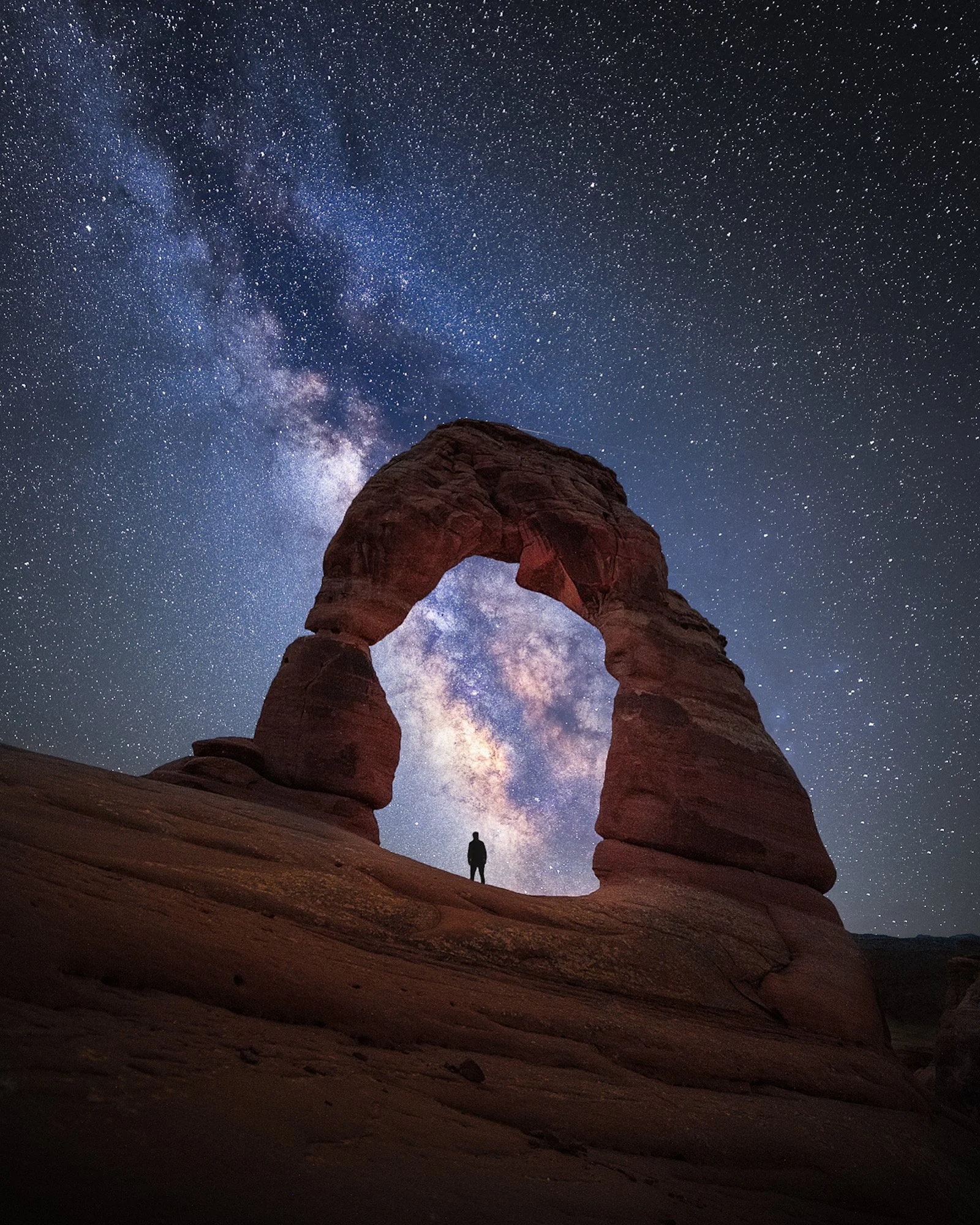 A night scene of Delicate Arch with a person standing underneath, stars and the Milky Way galaxy visible in the sky.