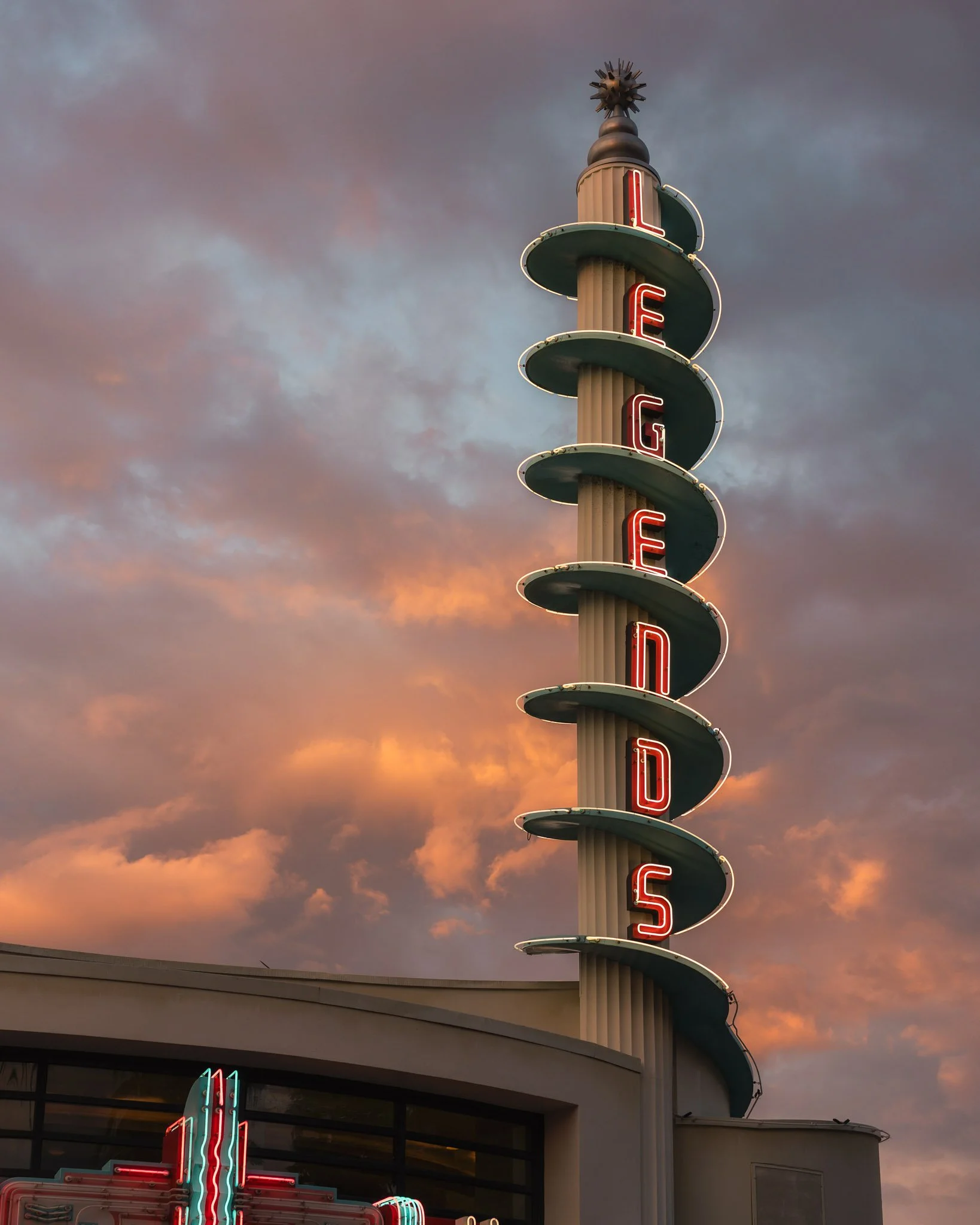 A vintage neon sign spelling 'LIGHTED' vertically, with each letter on a separate circular platform, set against a cloudy sunset sky.