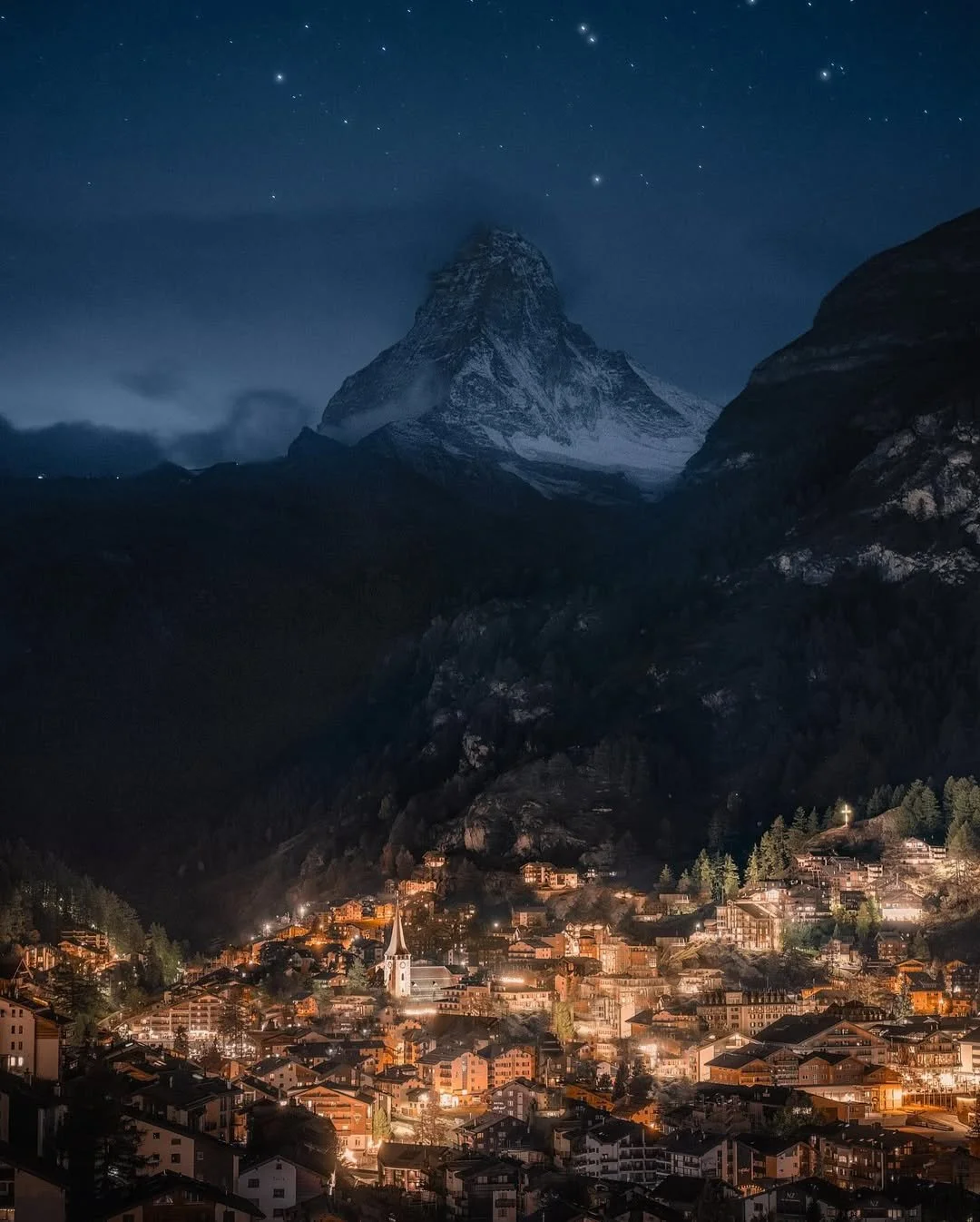 Nighttime view of a mountain with snow, stars above, and a lit town at the mountain's base