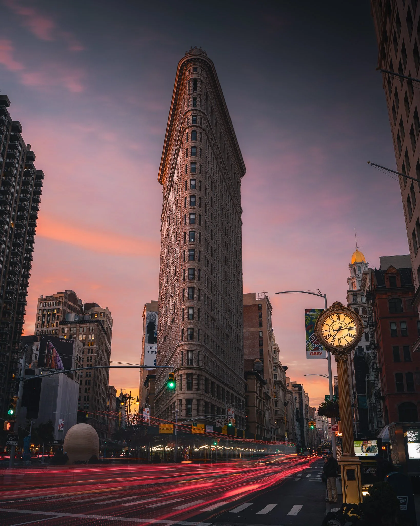 The iconic Flatiron Building in New York City during dusk, with city streets showing light trails from moving vehicles, a historic clock with gold accents, illuminated billboards, and surrounding buildings under a pink and purple sky.