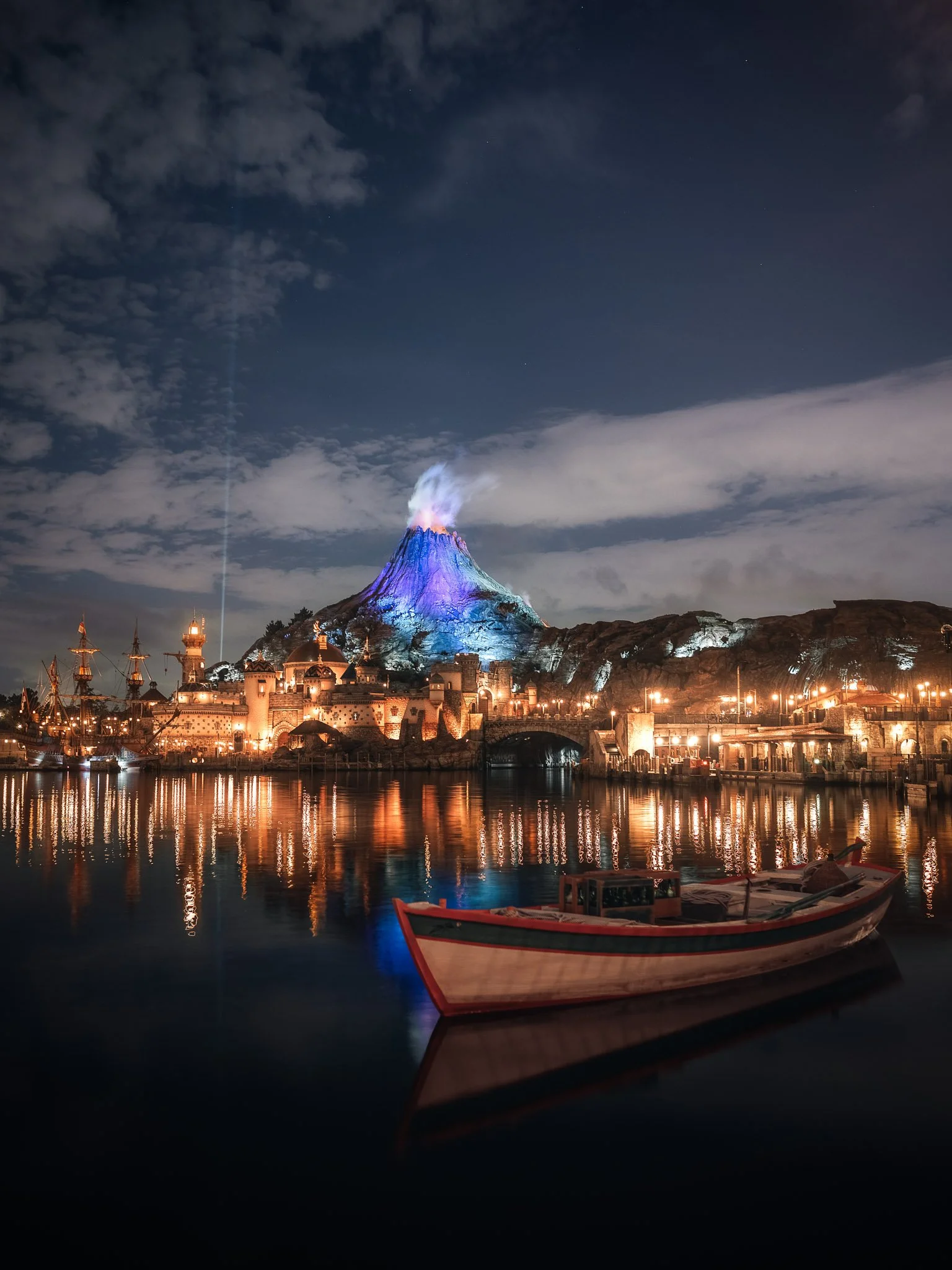 Nighttime scene at a harbor with a volcano erupting in the background, lit in blue and purple hues. The water reflects the lights and the erupting volcano. A boat is docked in the foreground.