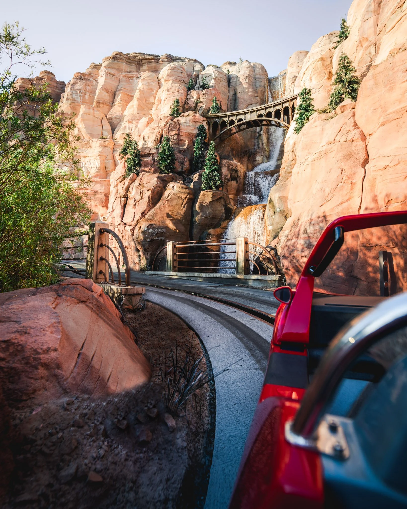 Red vehicle on mountain road with rock formations, trees, and a bridge in the background.