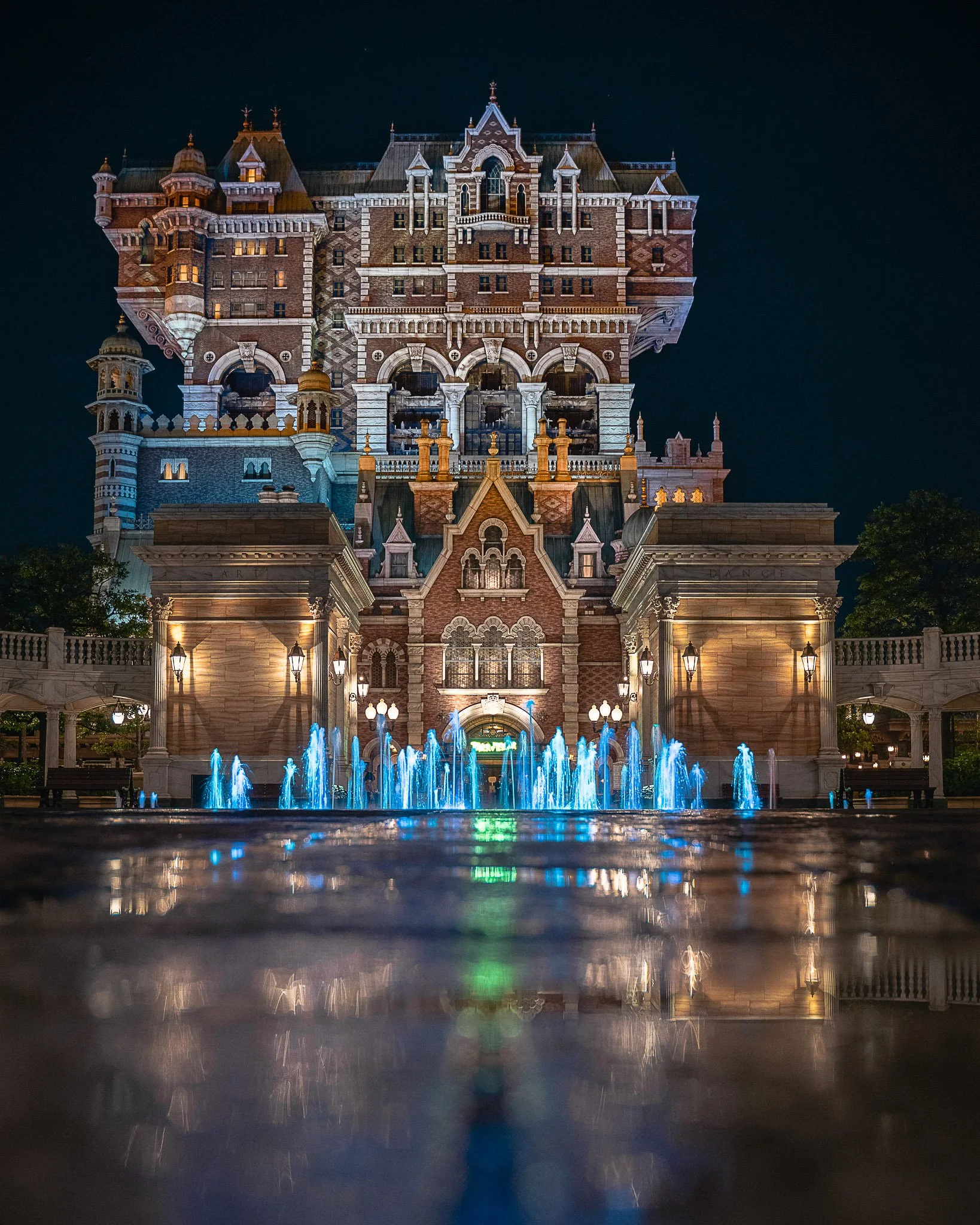 Night view of a grand, castle-like building with illuminated windows and a fountain with blue lights in the foreground.