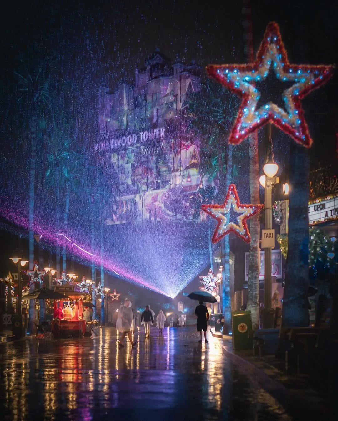 Night scene of Hollywood Boulevard with illuminated star-shaped decorations, wet pavement reflecting colorful lights, and a projection on the building facade.