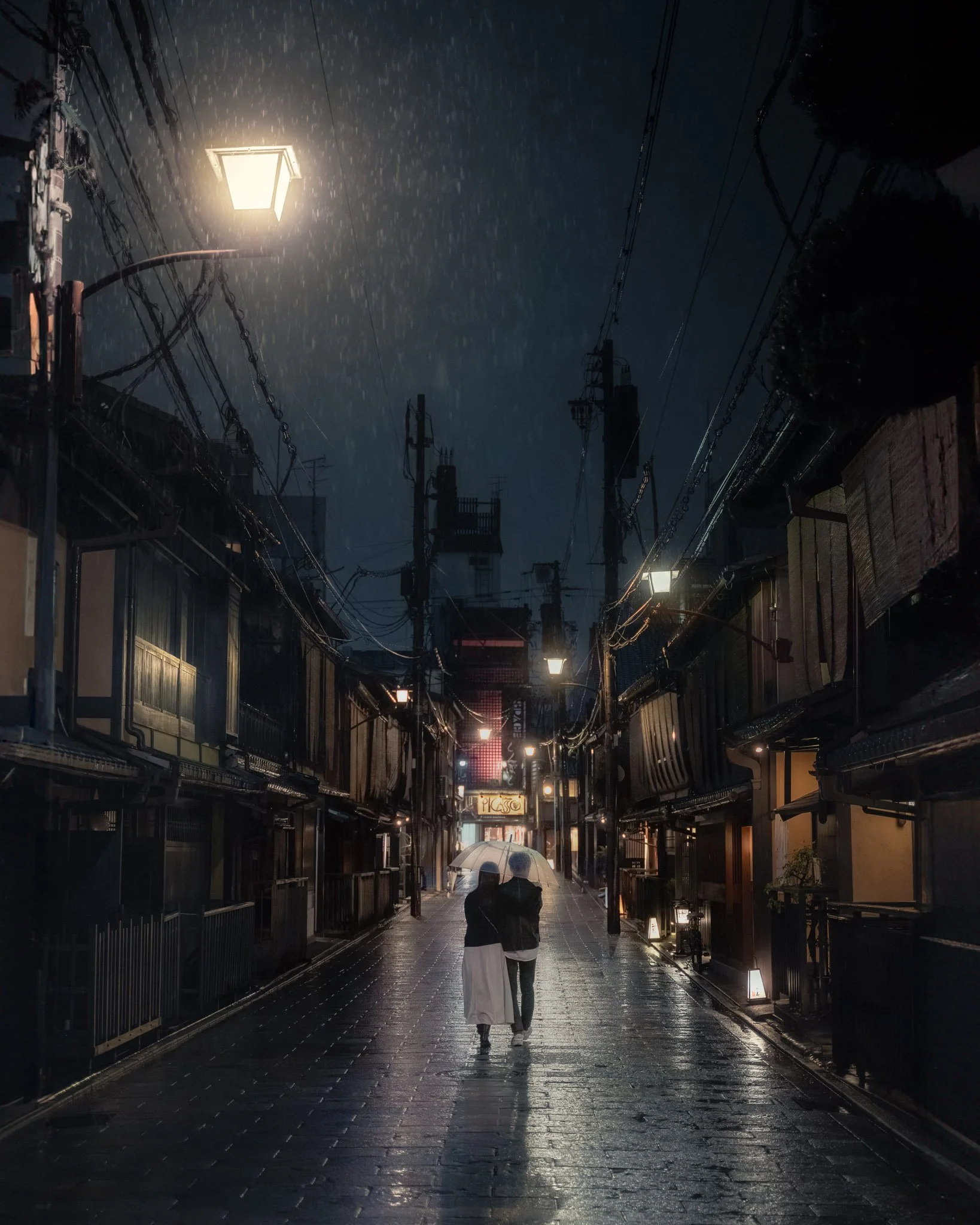 Nighttime scene of a rainy street with a couple walking under umbrellas. The street is wet and illuminated by streetlights, with power lines overhead and shopfronts lining both sides of the street.