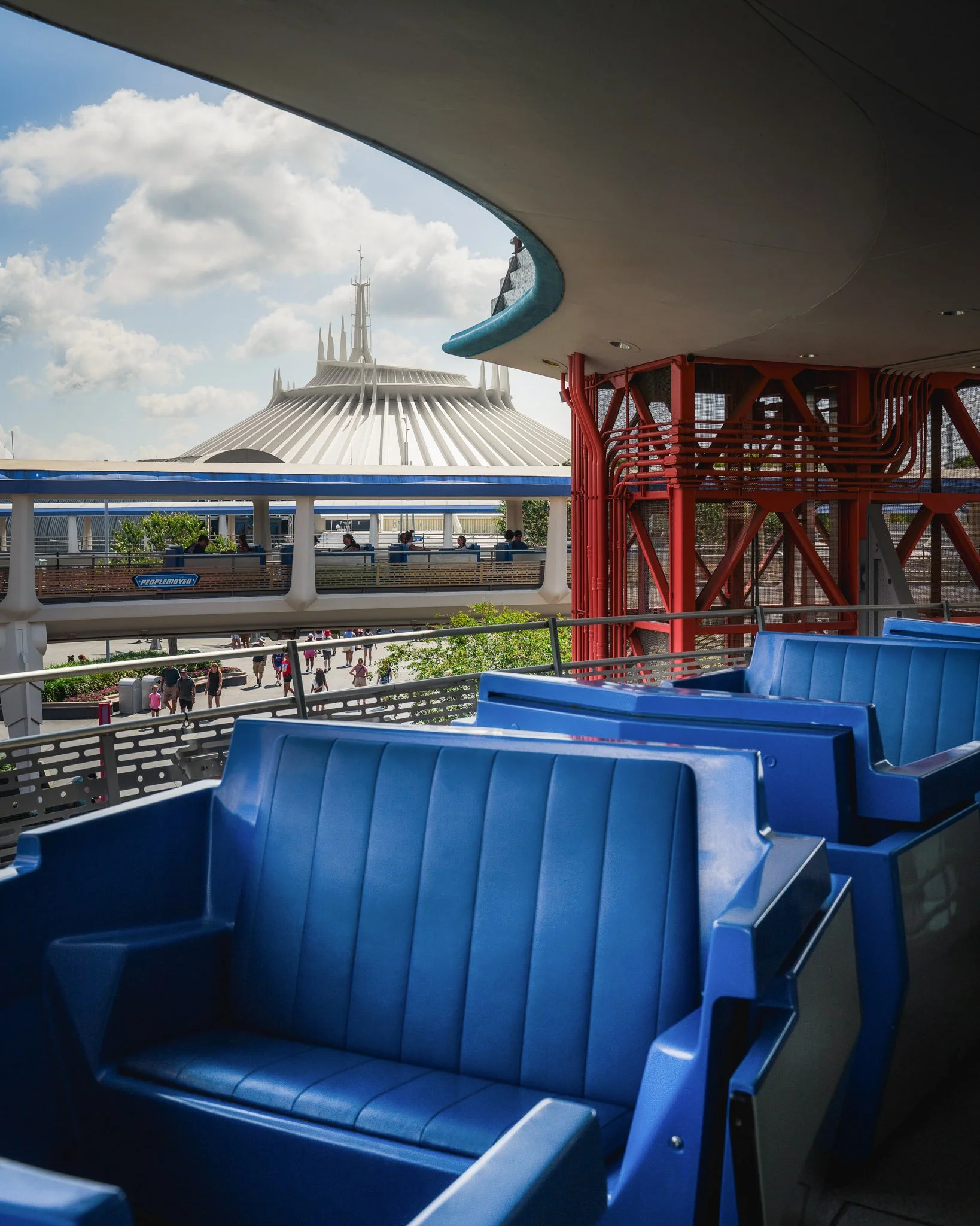 Blue empty seats on a transportation ride, with a view of futuristic structures and a partly cloudy sky in the background.