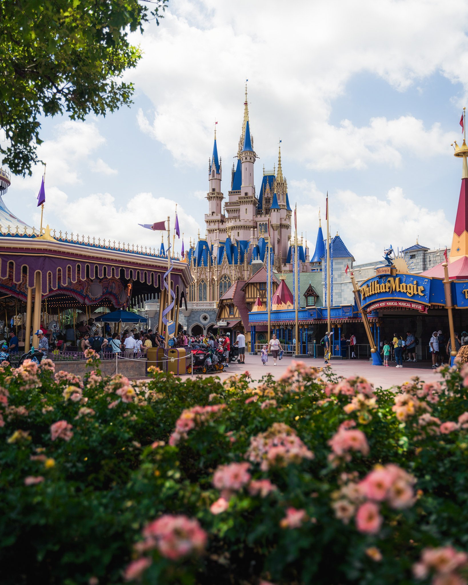Main view of Cinderella's Castle at a Disney theme park with people in line and pink flowers in the foreground under a cloudy sky.