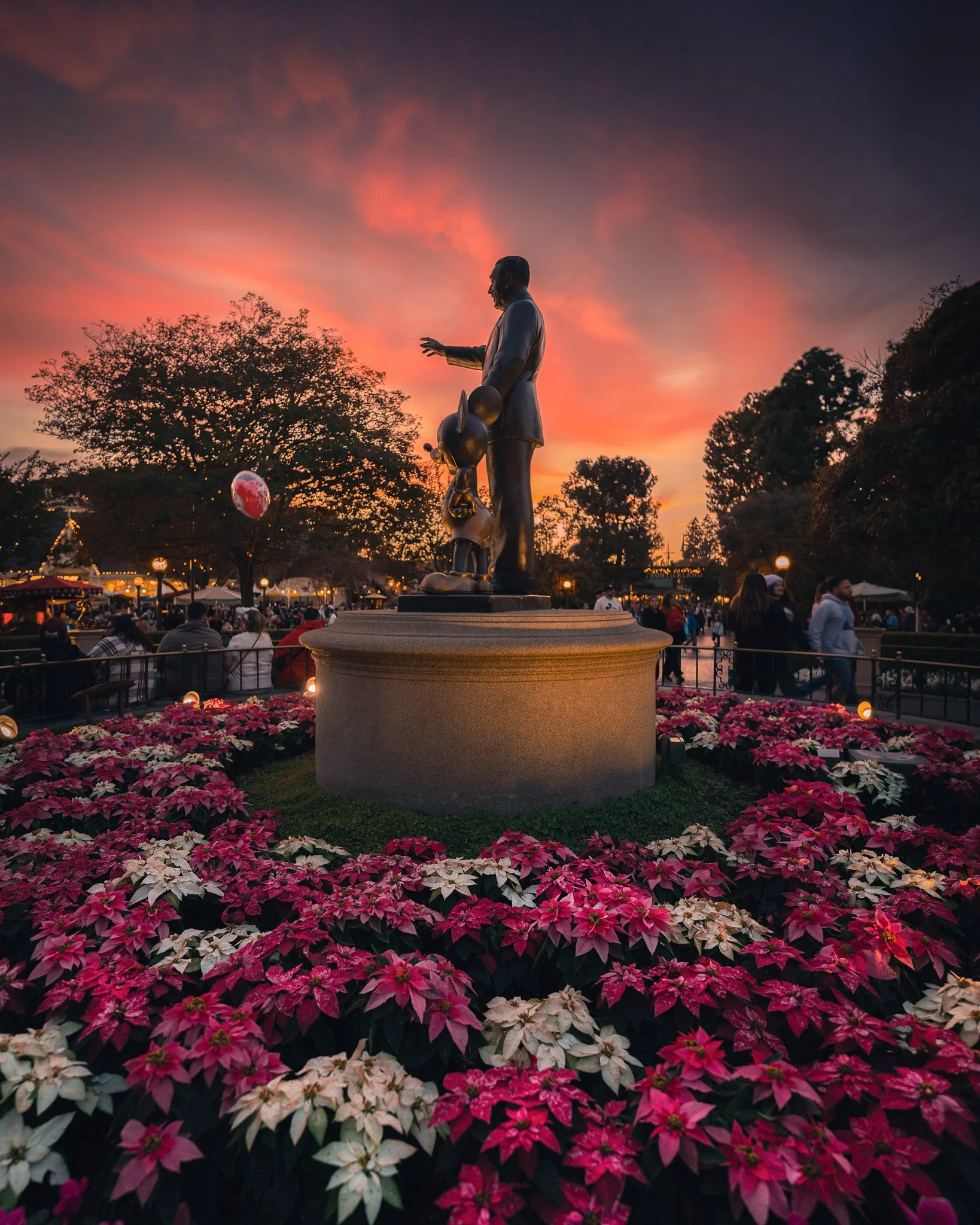 Statue of a man with a small girl at sunset, surrounded by pink and white flowers in a park.