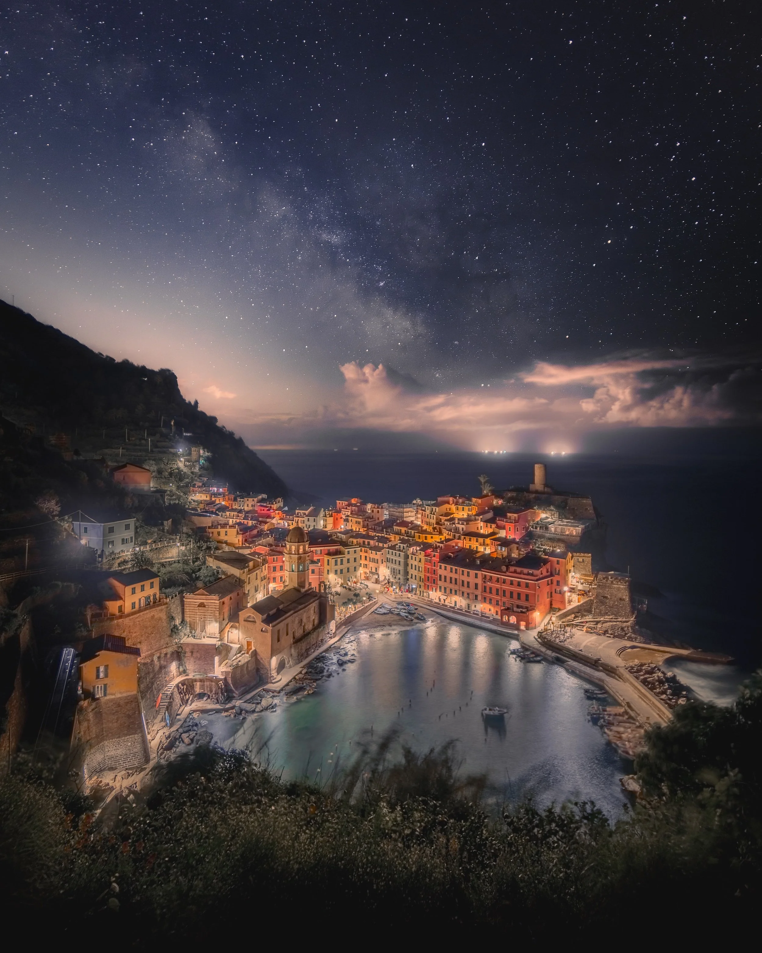 Nighttime view of a coastal town with colorful buildings, a harbor, and a hill with trees on the left, under a starry sky with the Milky Way.
