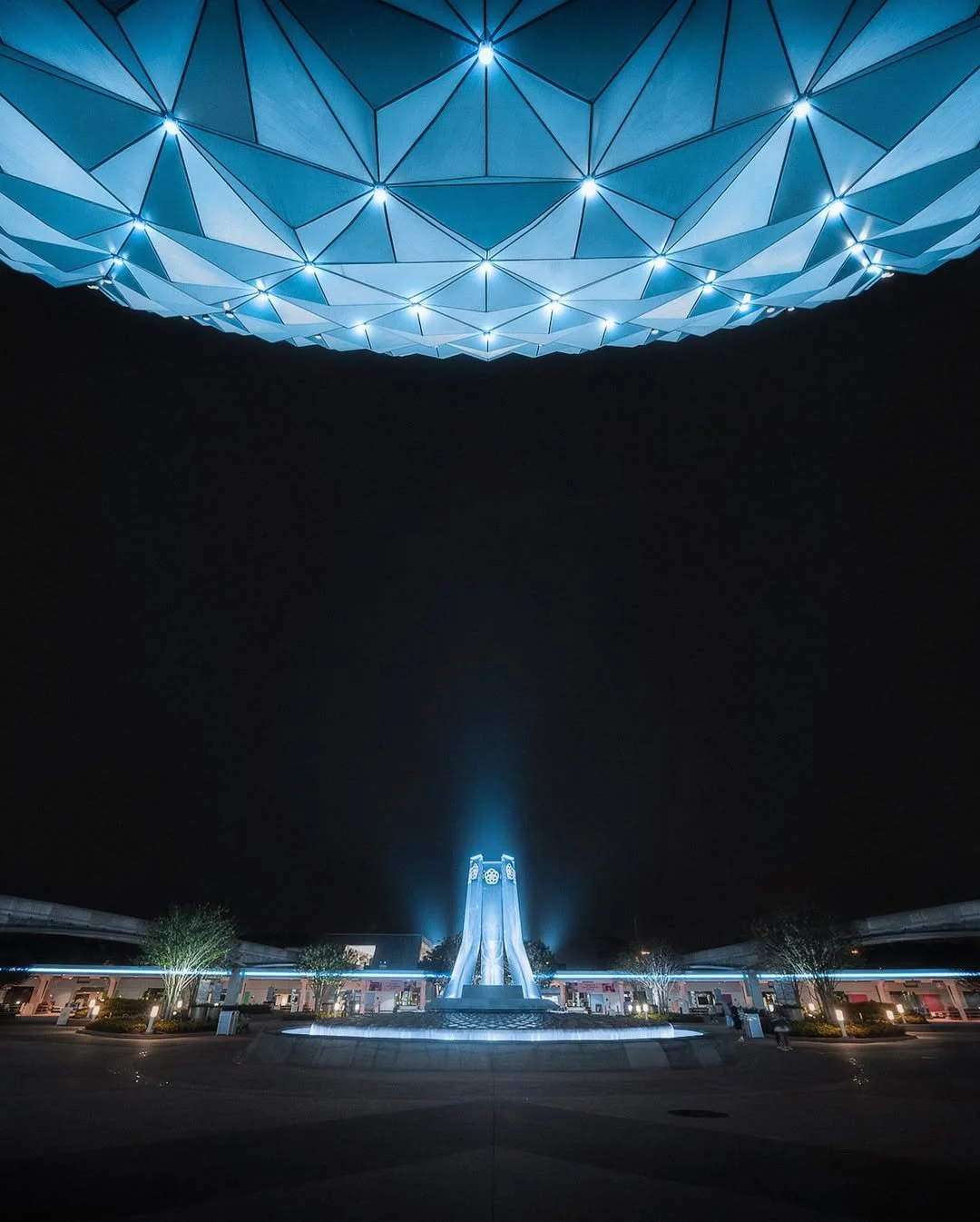 Night view of a futuristic illuminated building and sculpture with a large, geometric, glowing canopy overhead, possibly a landmark or monument.