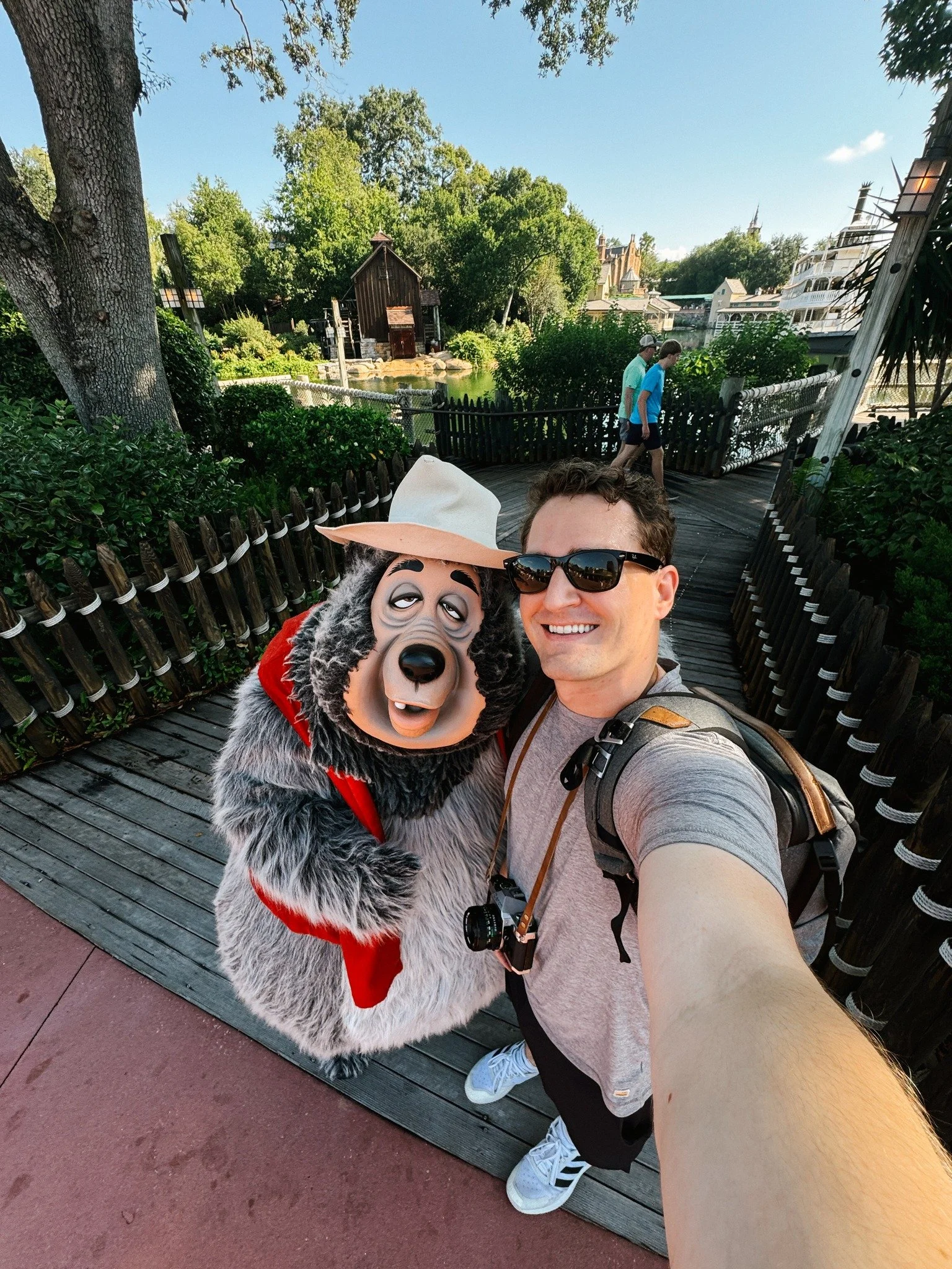 A man taking a selfie with a person in a costume of Baloo the bear from Disney's Jungle Book at a theme park. The background shows a pond, trees, and a wooden bridge.