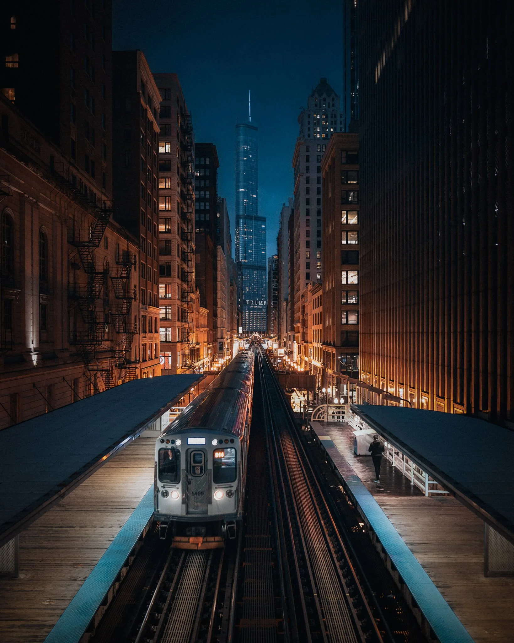 Night scene at a subway station in New York City with an approaching train, tall buildings, and One World Trade Center illuminated in the background.