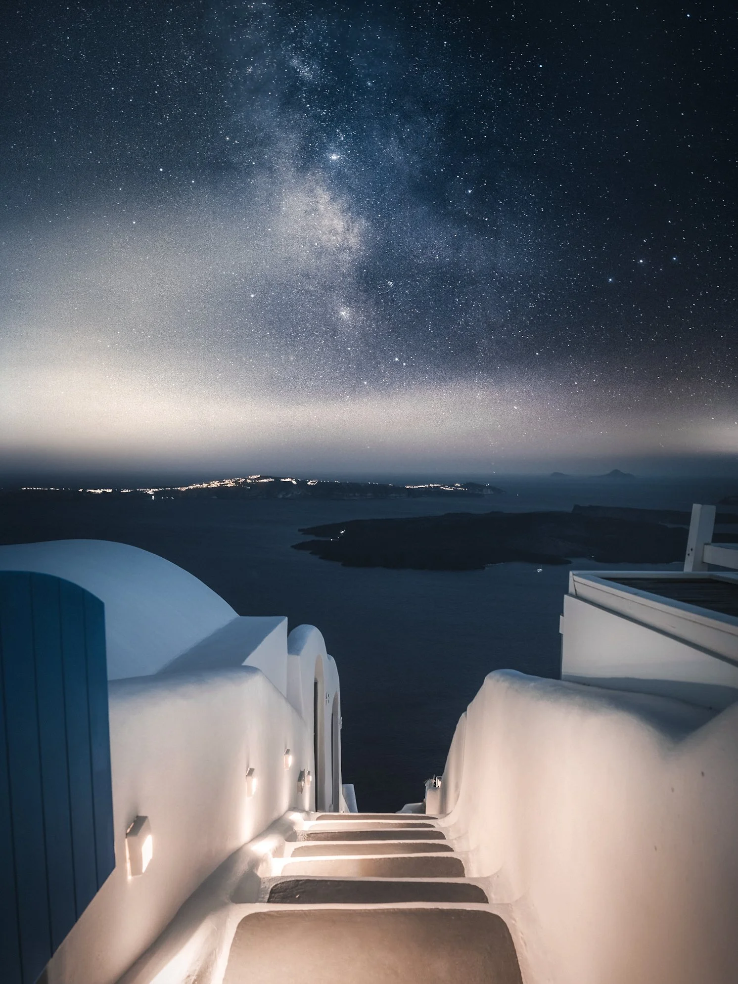 Whitewashed buildings with curved roofs and steps lead down to the water, overlooking a calm sea under a night sky filled with stars and the Milky Way galaxy.