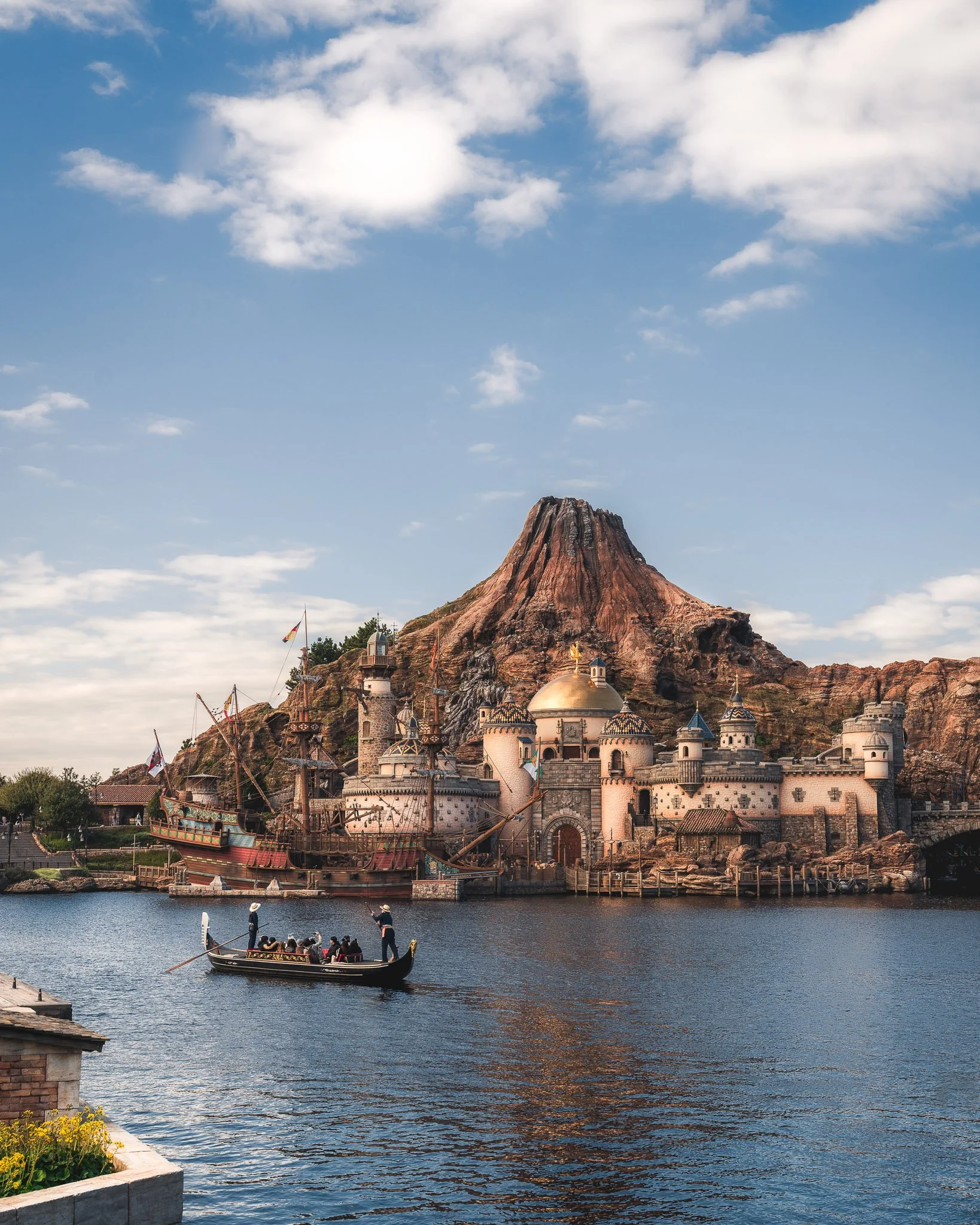 A pirate ship themed boat on a body of water in front a castle like building built into a rocky mountain, with a large ship on top, under a partly cloudy sky.