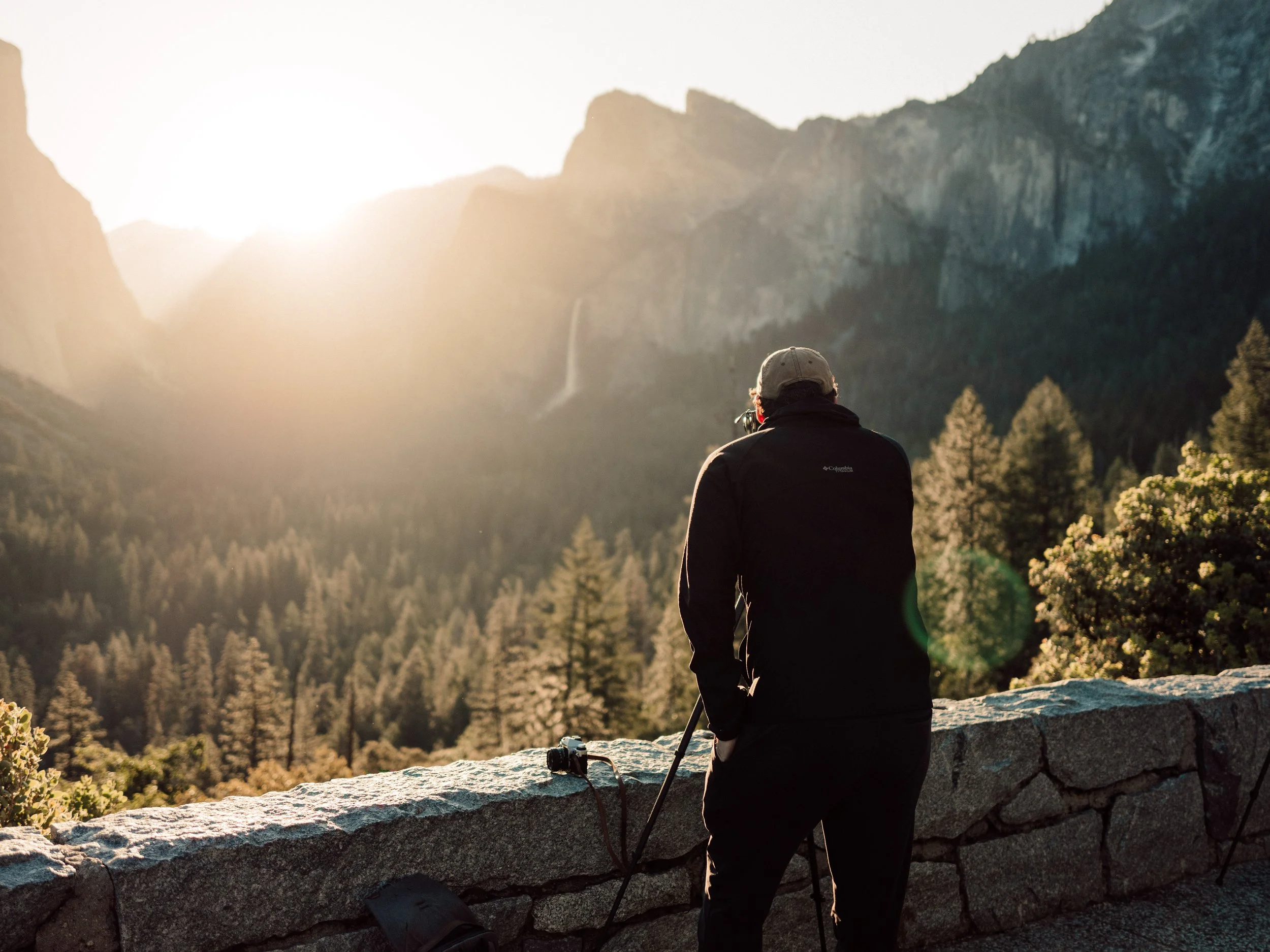 A person standing on a stone ledge, looking at a mountain landscape during sunset, camera resting on the ledge.