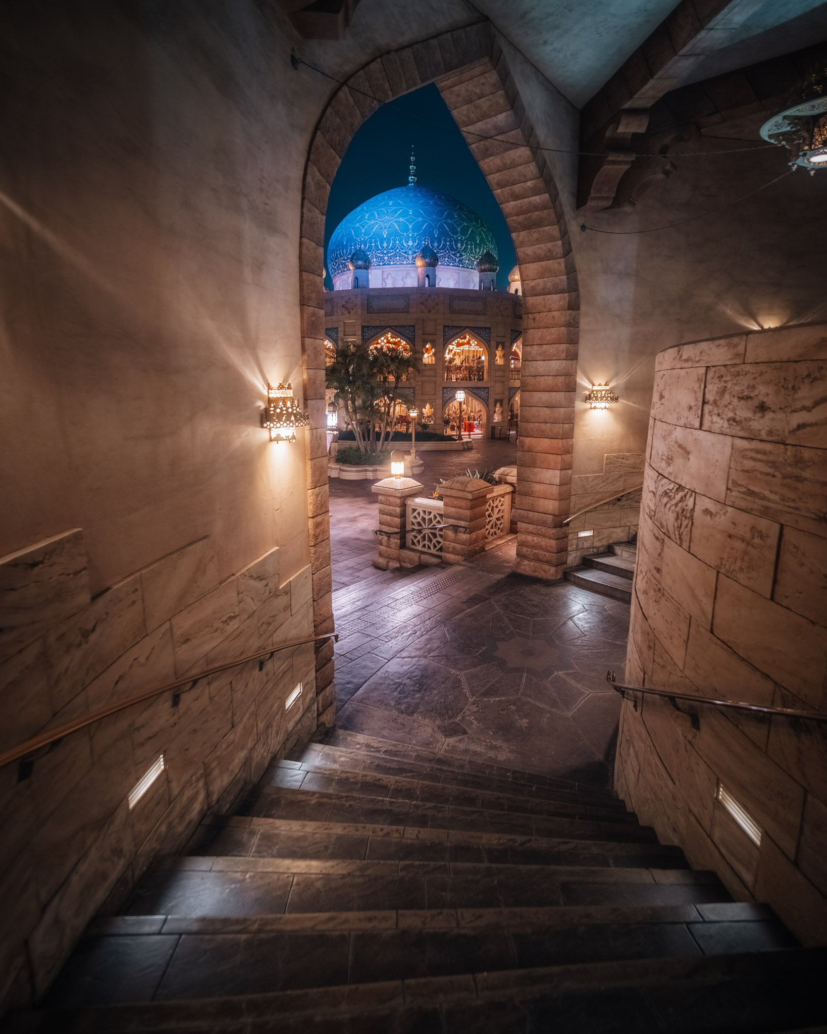 Night view of a mosque with a large illuminated blue dome and multiple minarets, seen through an arched stone passageway with staircase and wall-mounted lights.