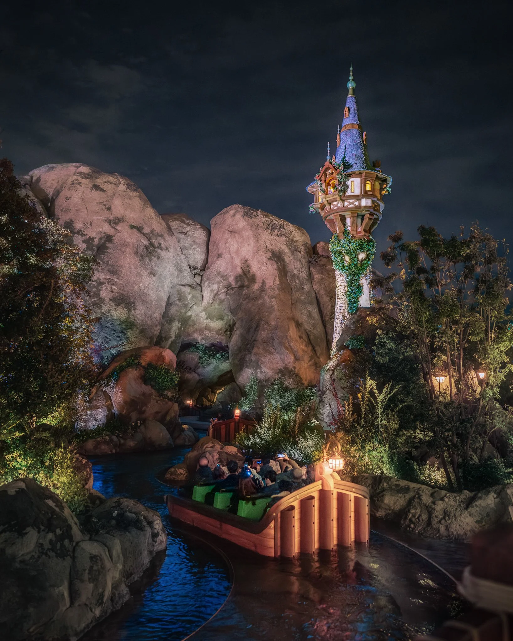Nighttime scene of a boat with visitors on a river, surrounded by large rocks and lush trees, with a fairy tale tower featuring a pointed, blue-shingled roof illuminated in the background.