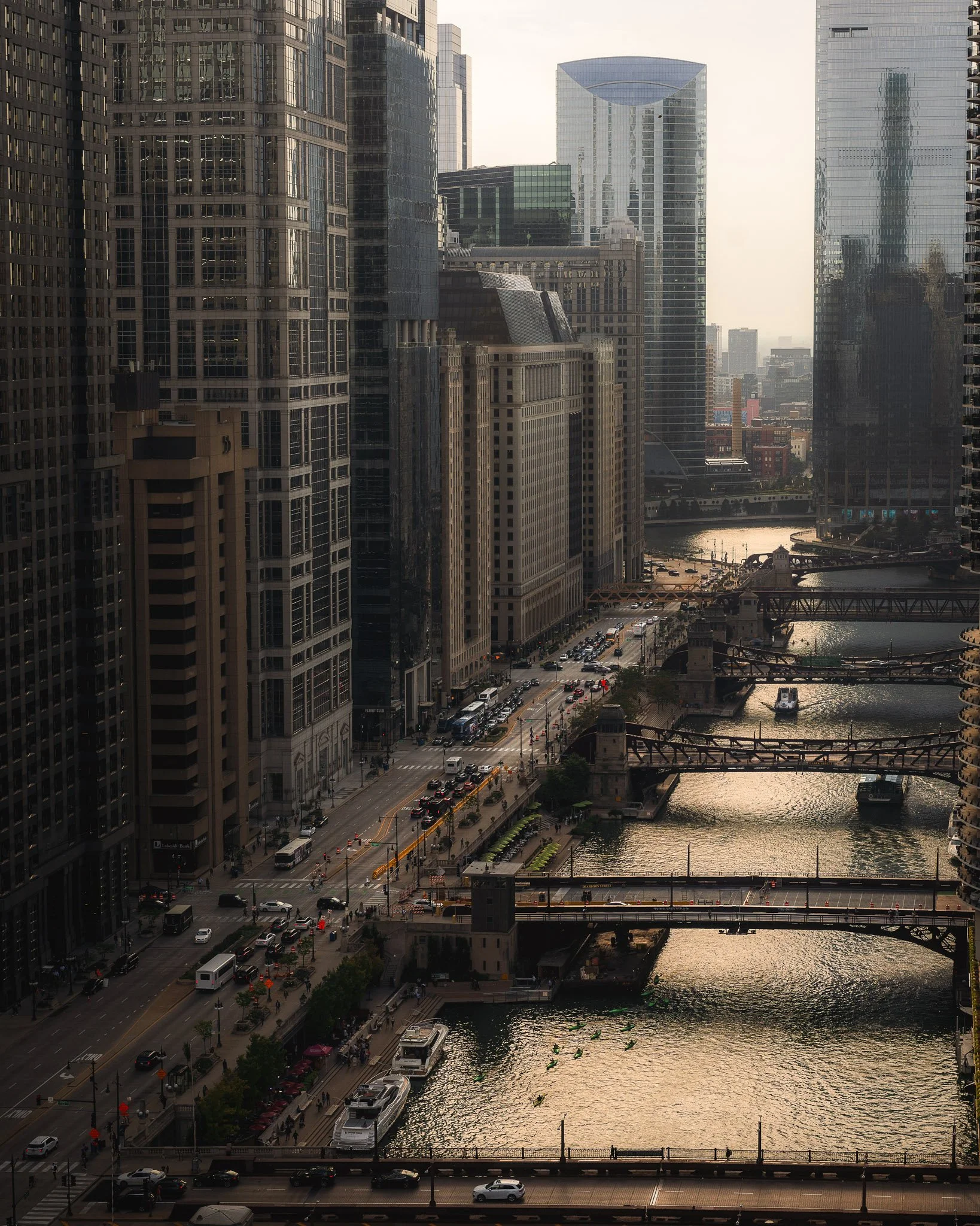 A cityscape with tall modern skyscrapers along a river, with bridges and a busy street with cars, buses, and pedestrians.
