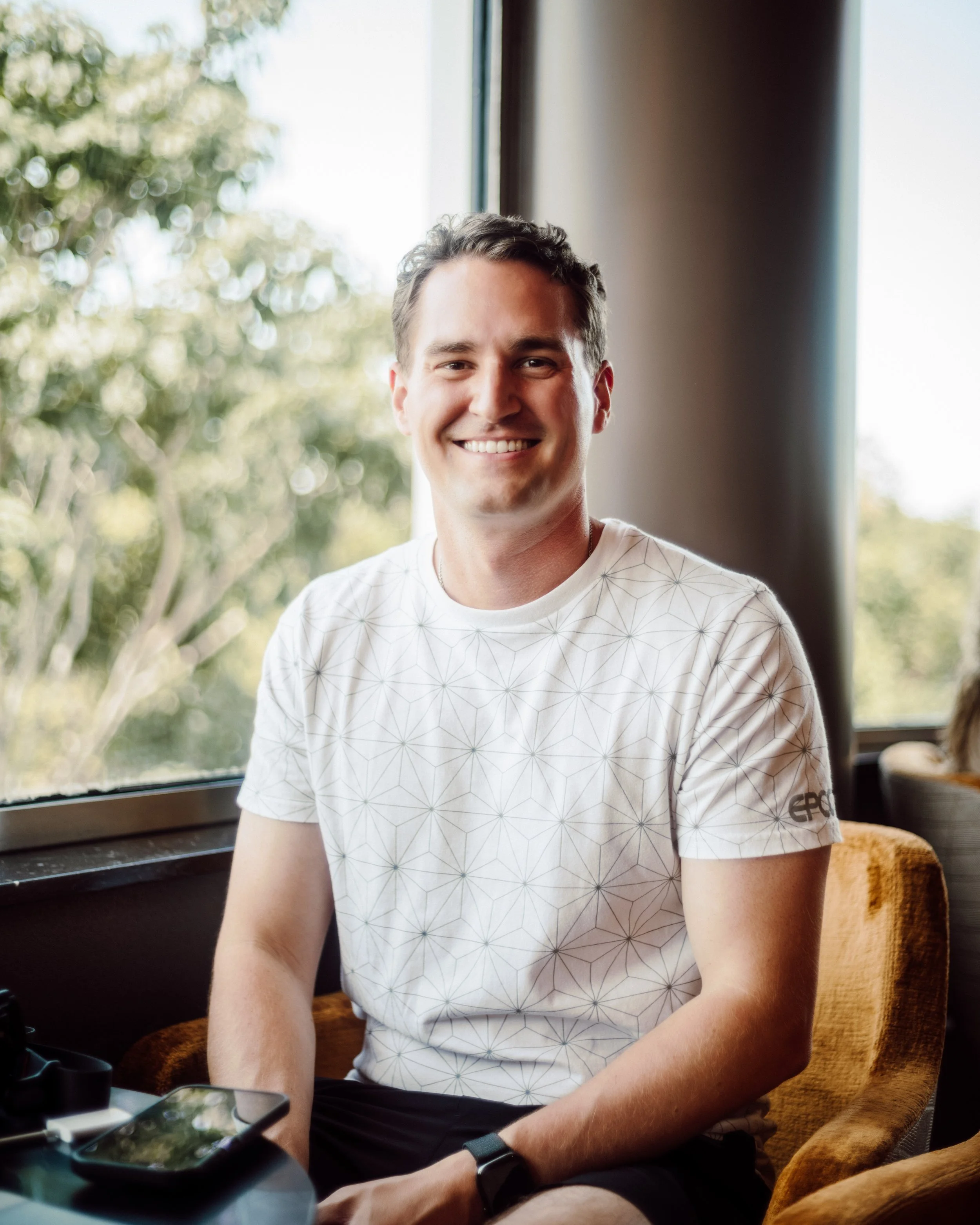 A smiling man with short dark hair, wearing a white geometric patterned t-shirt, sitting in a cozy indoor setting near a window with blurred trees outside.
