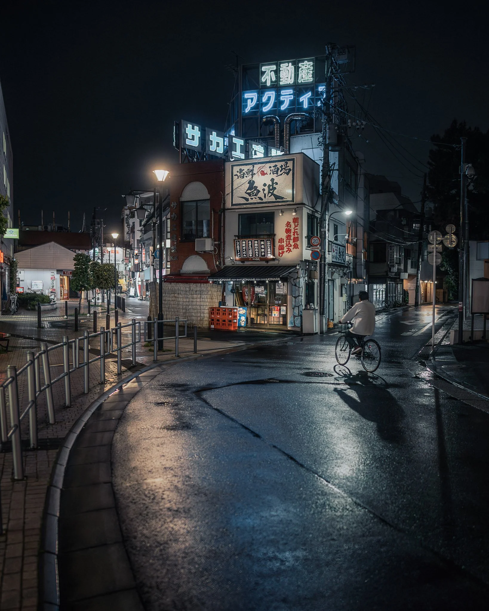 Nighttime urban street scene with illuminated signs in Japanese, a person riding a bicycle, wet pavement reflecting lights, streetlamps, and buildings.