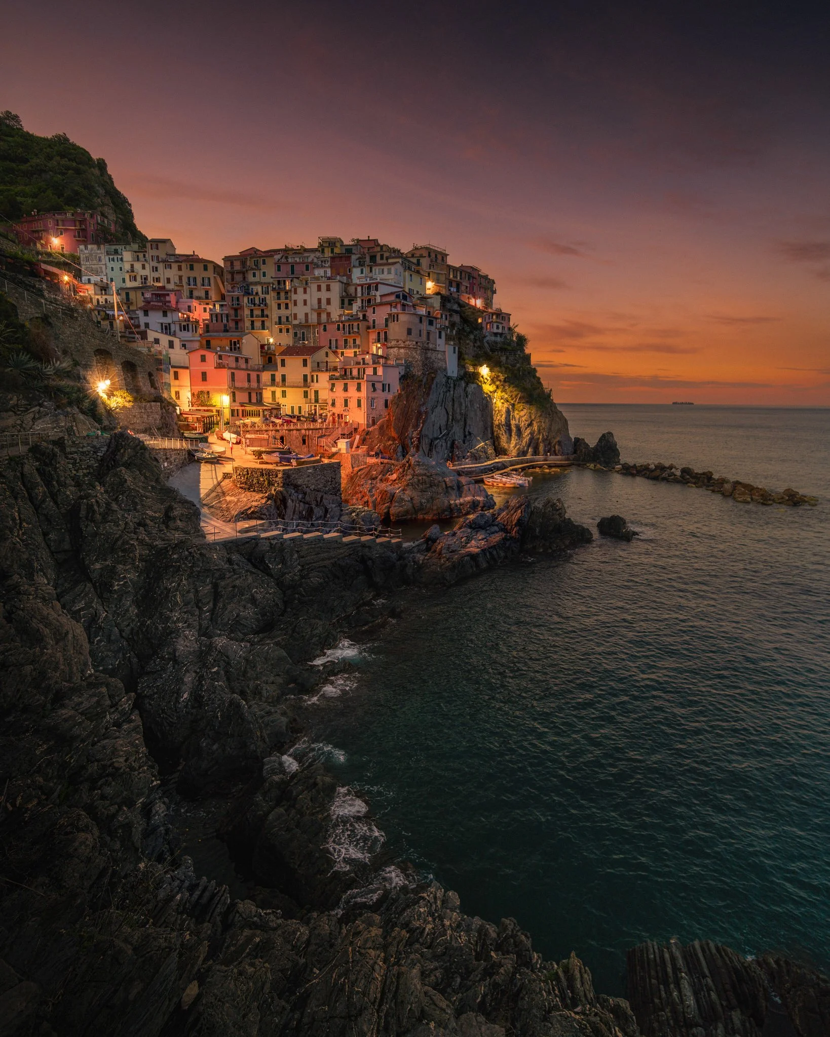 A coastal village at sunset with colorful buildings on a cliff overlooking the ocean, with boats docked near the rocky shoreline and a ship on the horizon.
