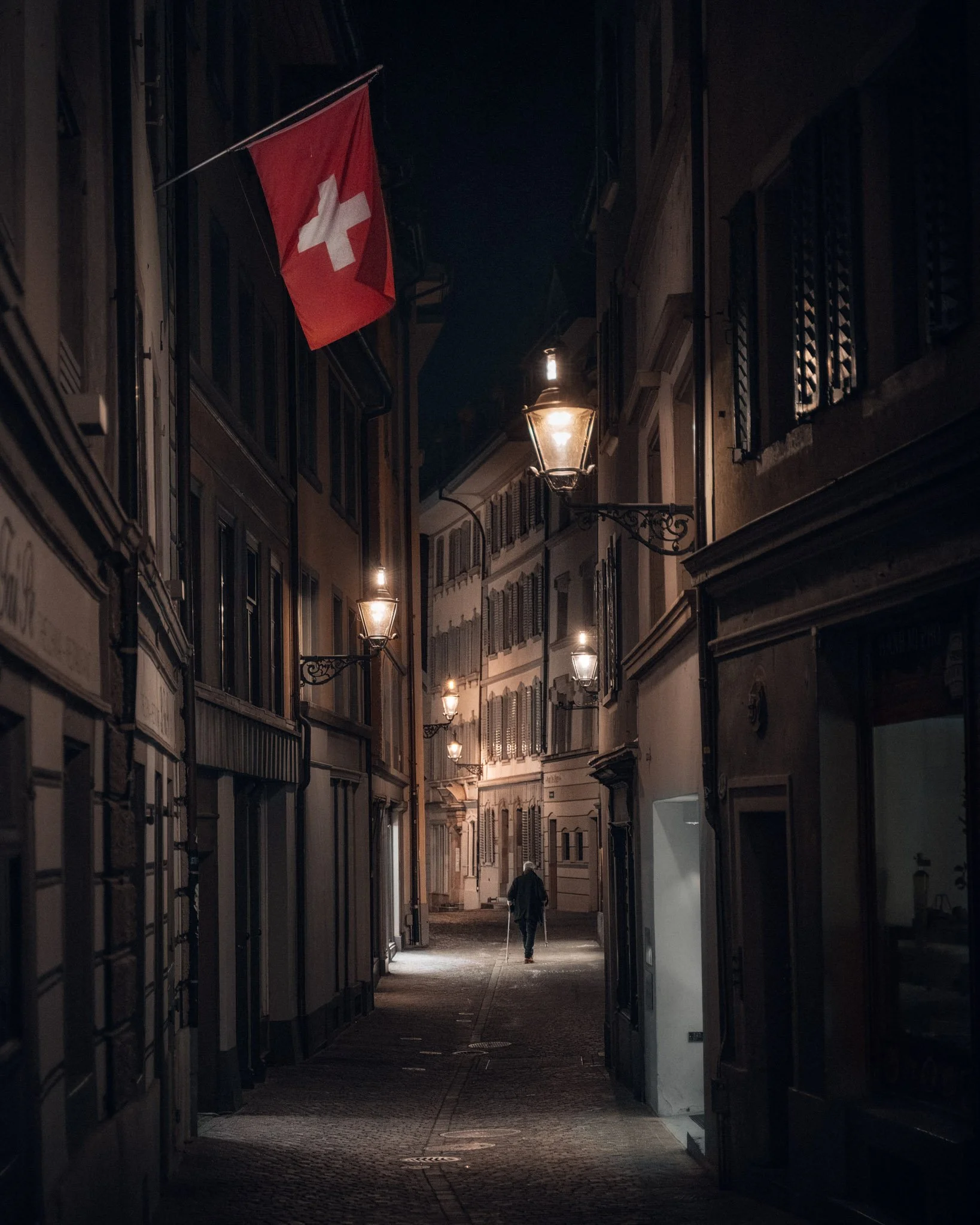 Nighttime street scene with a Swiss flag hanging on a building, illuminated street lamps, and a person walking down the alley.