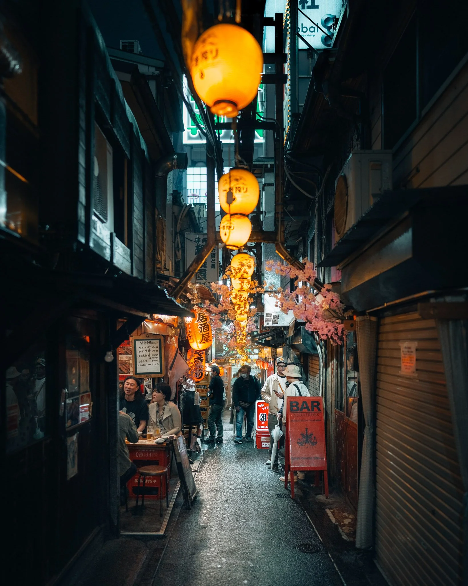 A narrow Japanese alleyway during rain with warm glowing lanterns hanging overhead, cherry blossom decorations, and people dining outside at restaurants.