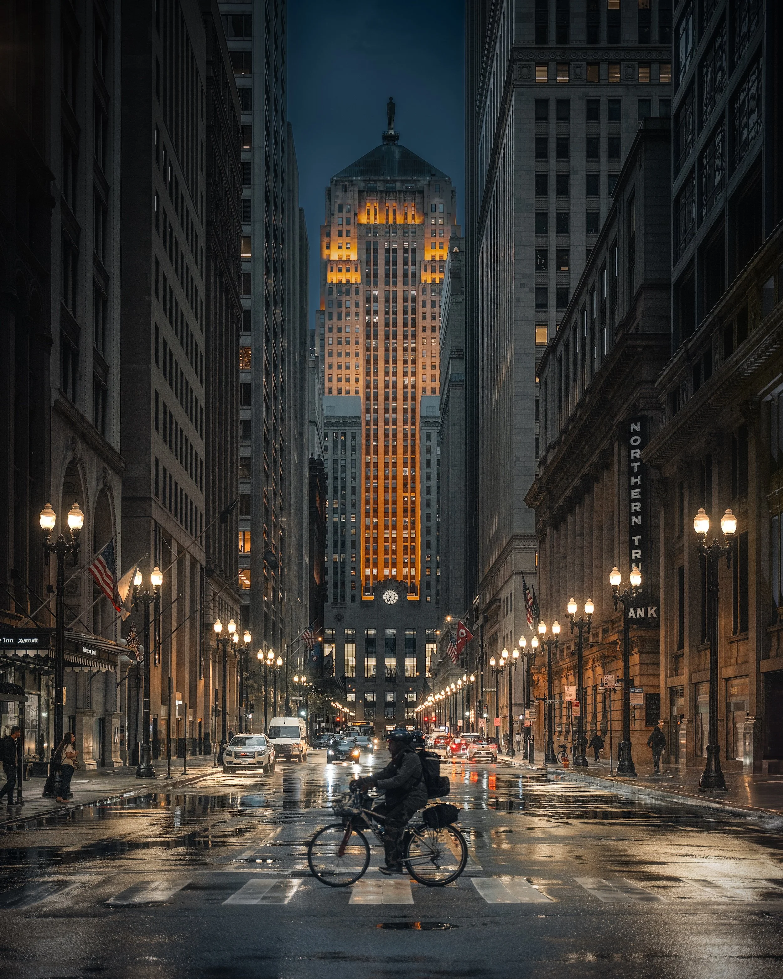 City street scene at dusk, wet pavement reflecting streetlights, tall buildings on either side, a person riding a bicycle in the foreground, cars and pedestrians around, illuminated building in the background with a clock at the bottom.