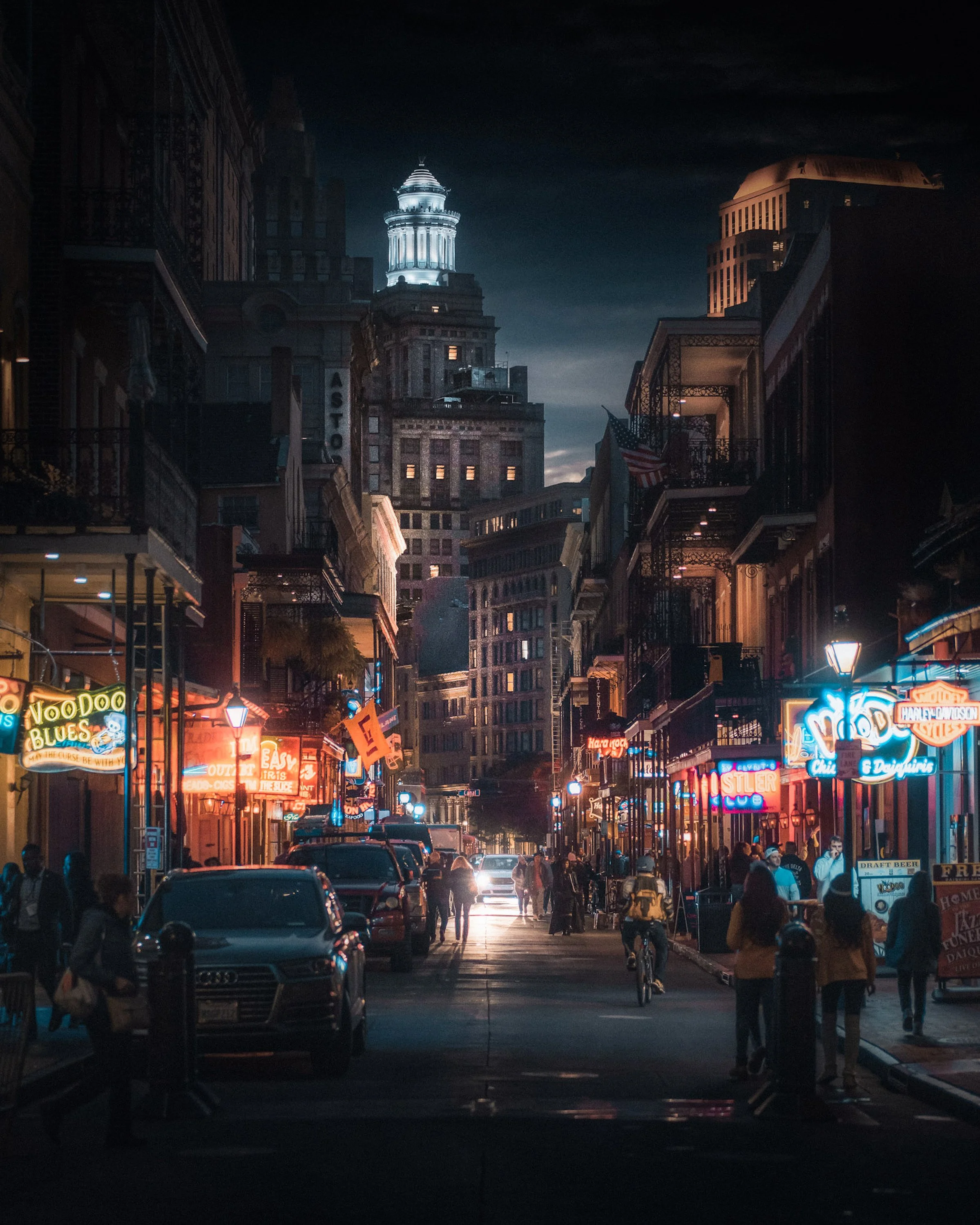 Nighttime urban street scene with lit neon signs, people walking, cars parked, and tall buildings including an illuminated clock tower in the background.