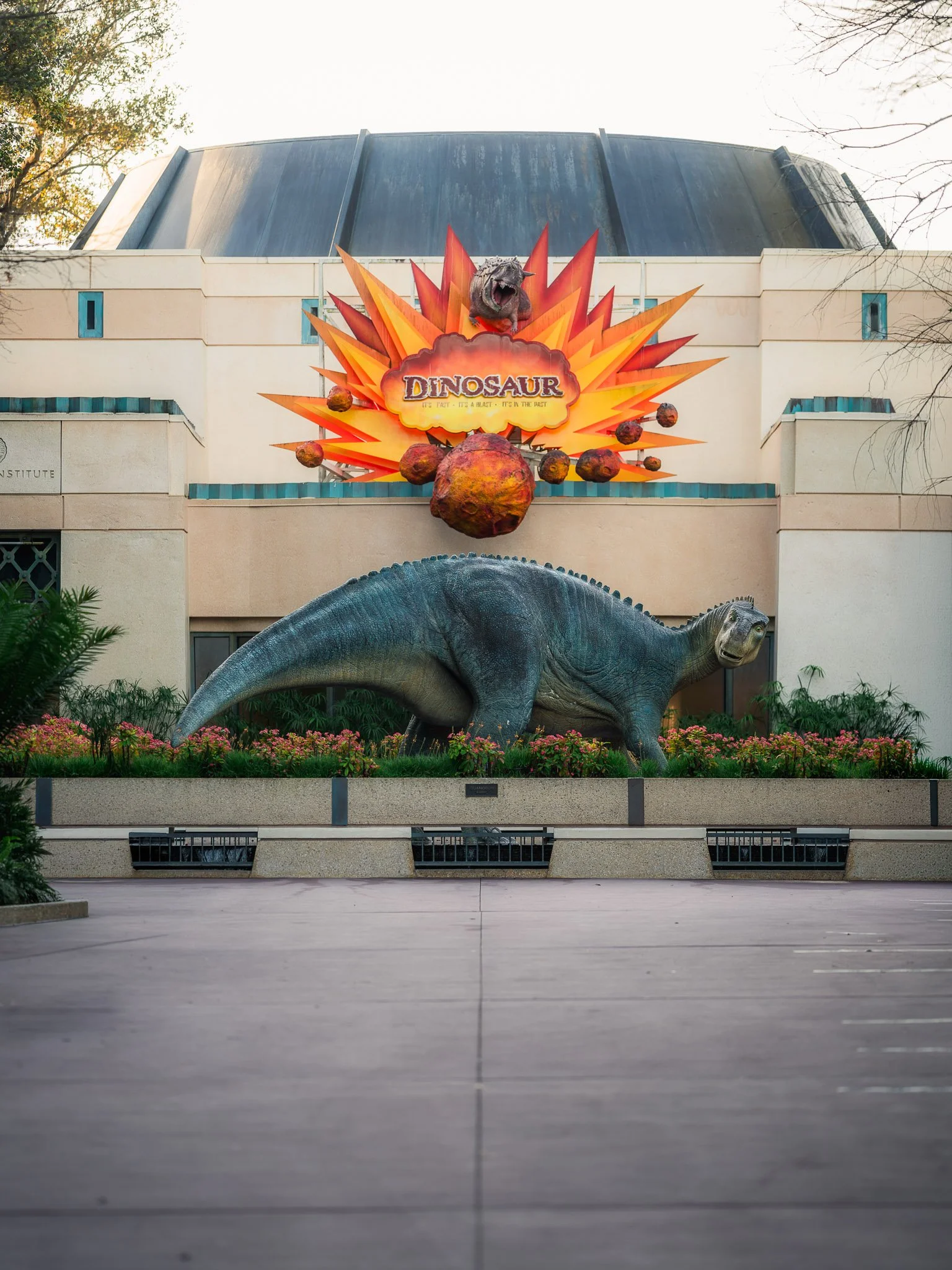 Museum building with a large dinosaur sculpture in front. The sculpture includes a dinosaur head and body, surrounded by a colorful, comic-like explosion with the word "DINOSAUR" in the center.