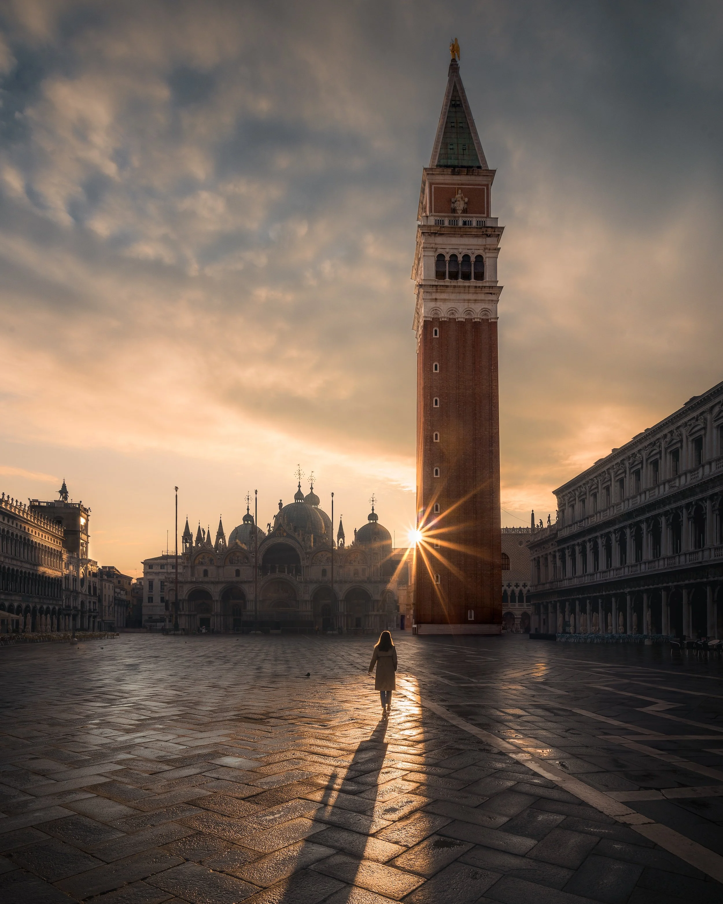 A person standing in Piazza San Marco in Venice at sunset, looking towards the famous clock tower, with the sky full of clouds and the sun casting long shadows on the cobblestone ground.