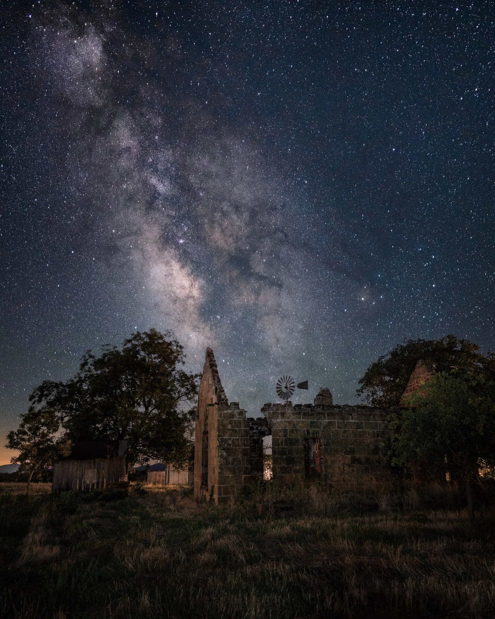 Night sky filled with stars and the Milky Way galaxy above an abandoned stone building with trees and grass in the foreground.