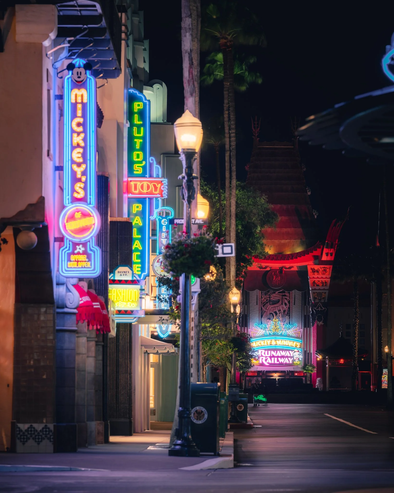 Nighttime street scene with colorful neon signs advertising Mickey's, Play Space, Toy, Munch, Mickey & Minnie Runaway Railway, and other shops. The street features arcade-style lighting, palm trees, decorative lamps, and empty sidewalks.