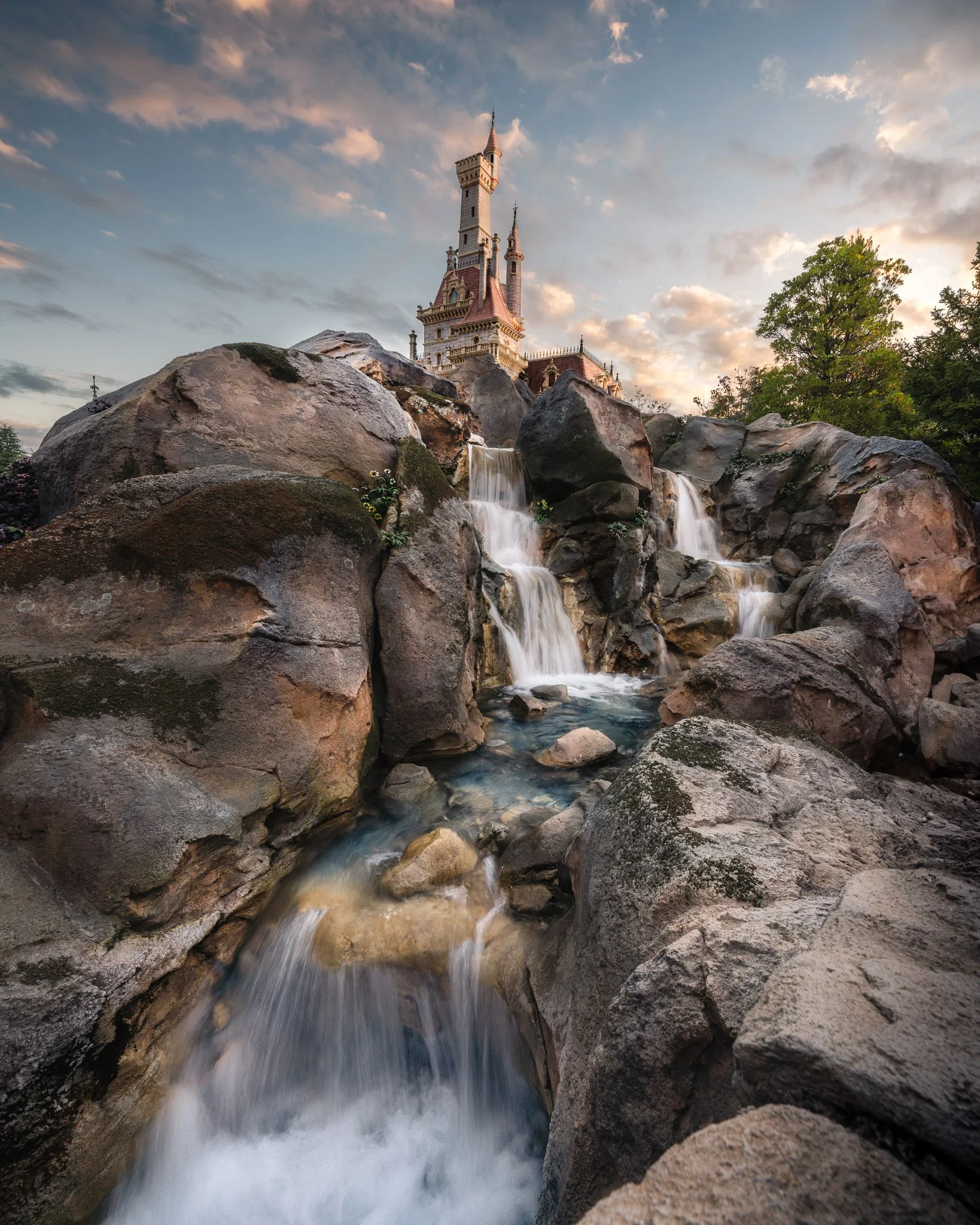 A castle with a tall pointed tower and smaller spires behind large rocks. A waterfall flows between the rocks, with a tree to the right, and a cloudy sky at sunset in the background.
