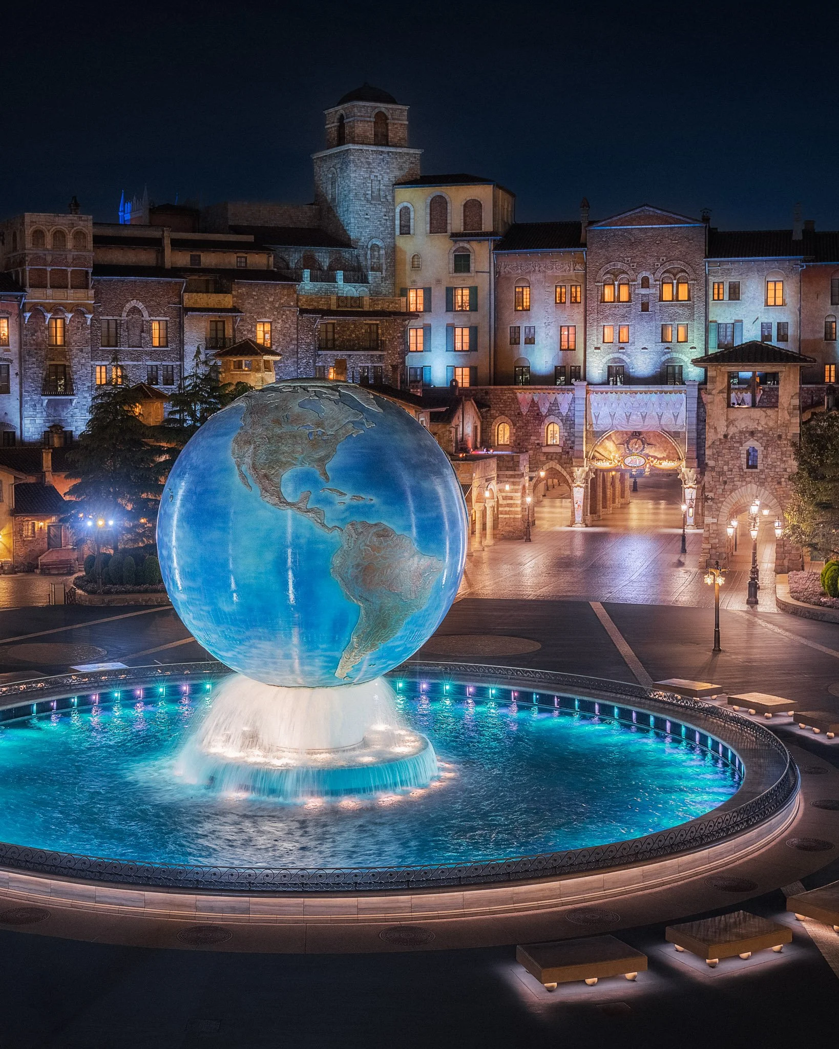A large illuminated globe fountain in front of a European-style building complex at night, with the fountain's water spray and glowing lights in the pool.