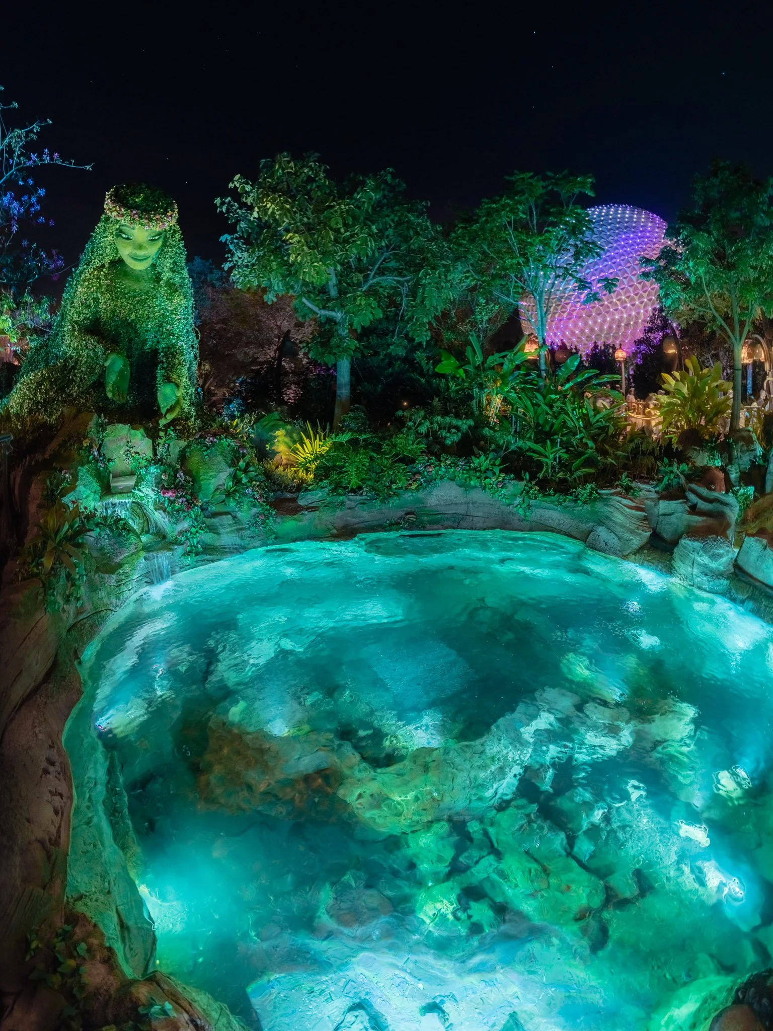 Nighttime scene of illuminated landscape featuring lush green trees, a glowing pond, a large spherical structure with purple lights, and a large statue of a woman covered in plants and flowers.