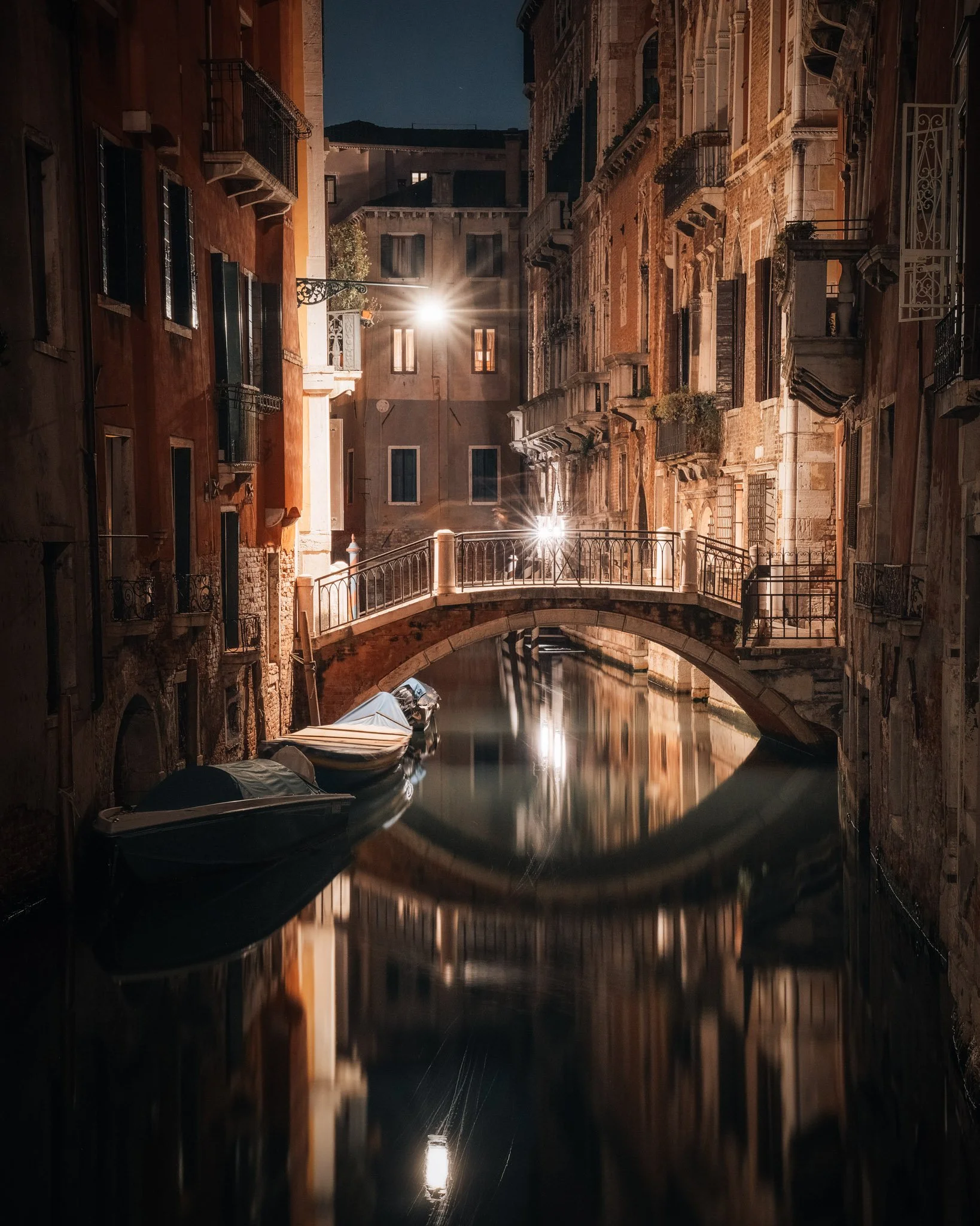 Nighttime view of a small canal with boats docked along the sides, illuminated by streetlights. Surrounding buildings have balconies and windows with warm lighting, creating a serene and picturesque scene.