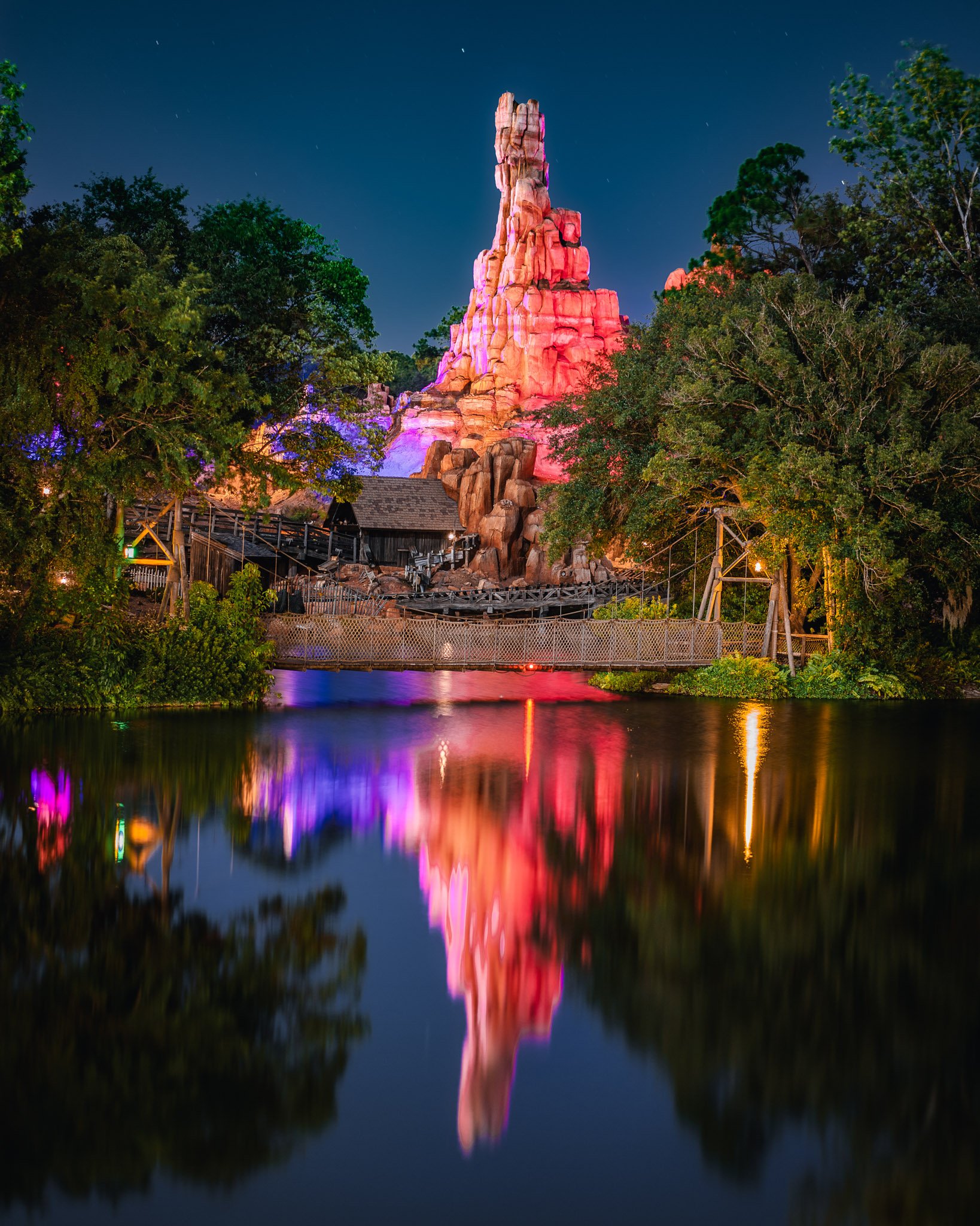 Night view of a tall, rocky formation illuminated with pink and purple lights, reflected in a calm body of water, surrounded by green trees.